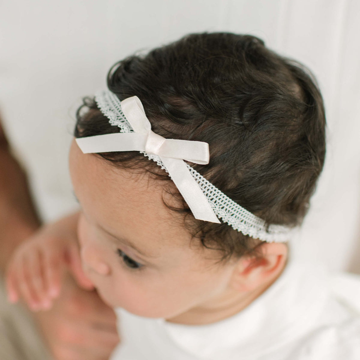 A close-up of a baby wearing an ivory lace headband with a pink satin bow, set against a soft white background.