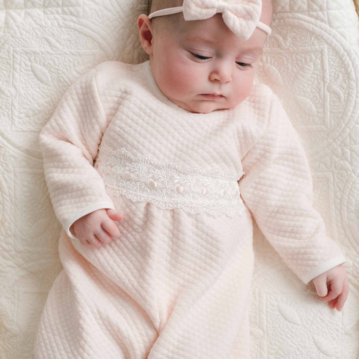 A sleeping newborn baby wearing a pale pink textured long-sleeve footless baby jumpsuit with a white lace band across the chest, along with a matching pink bow headband, lying on a white quilted blanket.