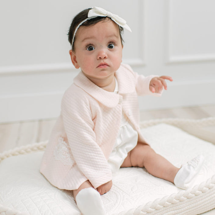 A baby girl wearing a pale pink textured cardigan, white romper, white booties, and a white bow headband, sitting on a white quilted blanket.
