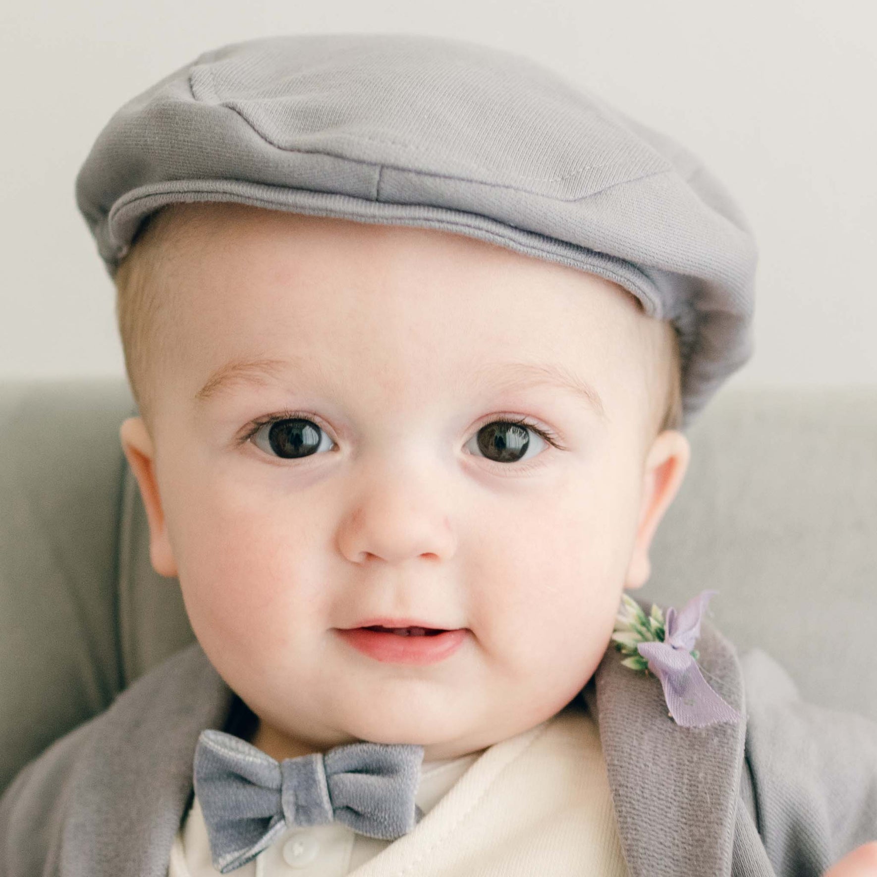 Baby boy sitting on a couch wearing the Ezra 4-Piece Suit with matching Newsboy Cap, heather velvet bow tie and boutonniere