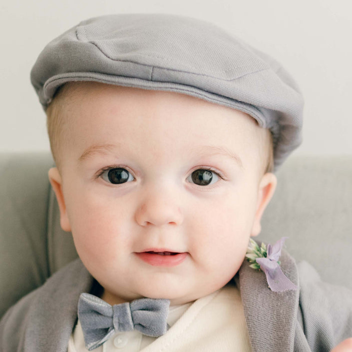 Baby boy sitting on a couch wearing the Ezra 4-Piece Suit with matching Newsboy Cap, heather velvet bow tie and boutonniere