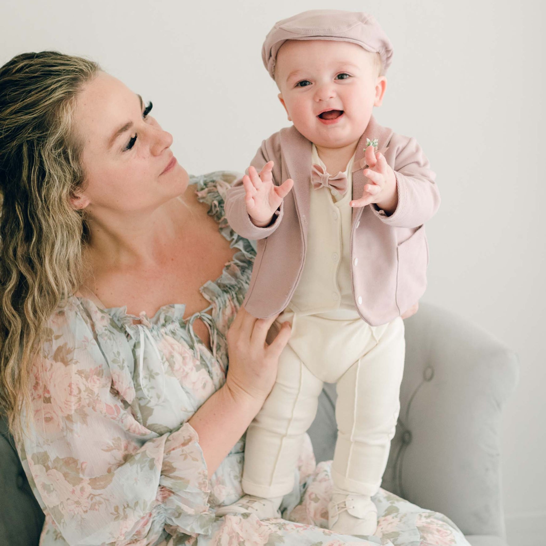 Baby boy standing on his mother's lap. He is wearing the Ezra 4-Piece Mauve Suit with matching Newsboy Cap, mauve velvet bow tie and boutonniere
