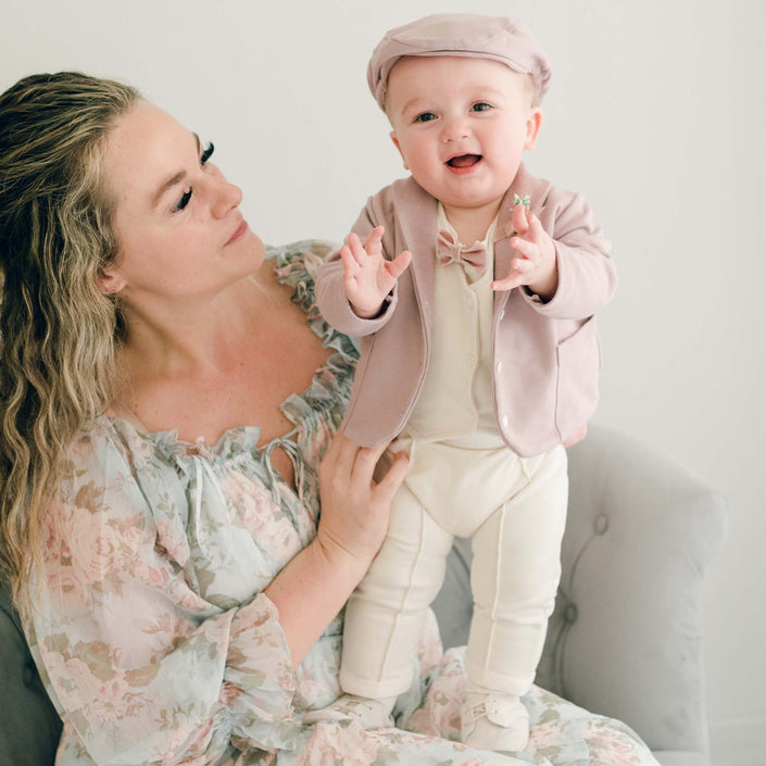 Baby boy standing on his mother's lap. He is wearing the Ezra 4-Piece Mauve Suit with matching Newsboy Cap, mauve velvet bow tie and boutonniere