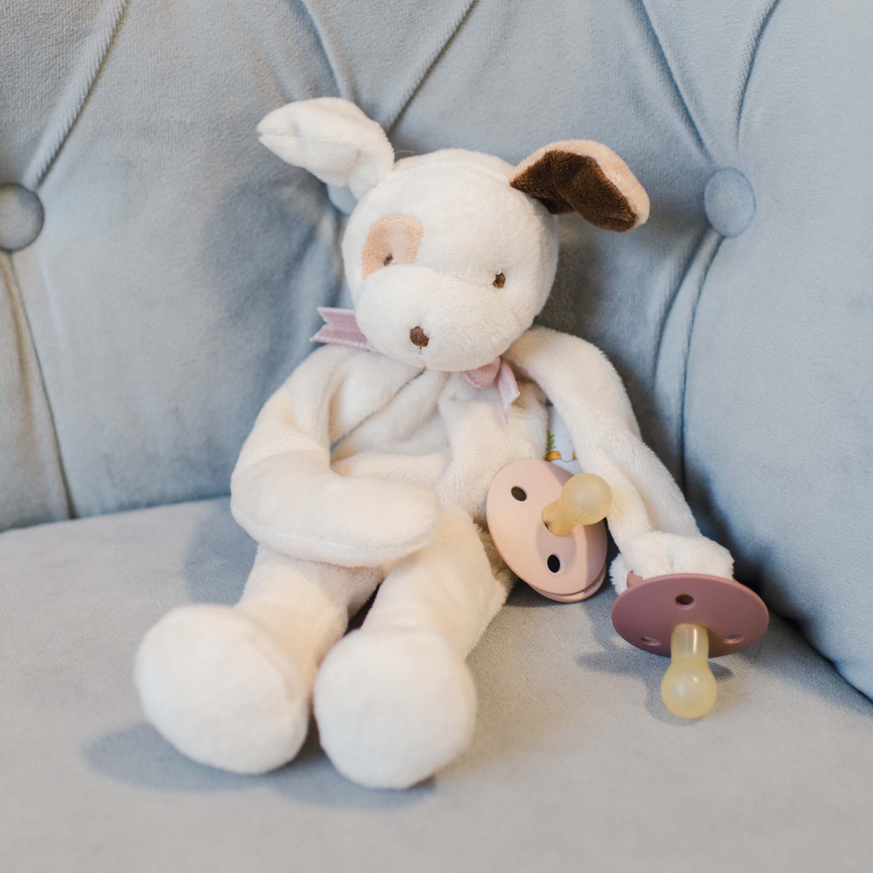 The Ezra Mauve Silly Puppy Buddy stuffed animal sitting on a couch. This stuffed animal serves as a pacifier holder