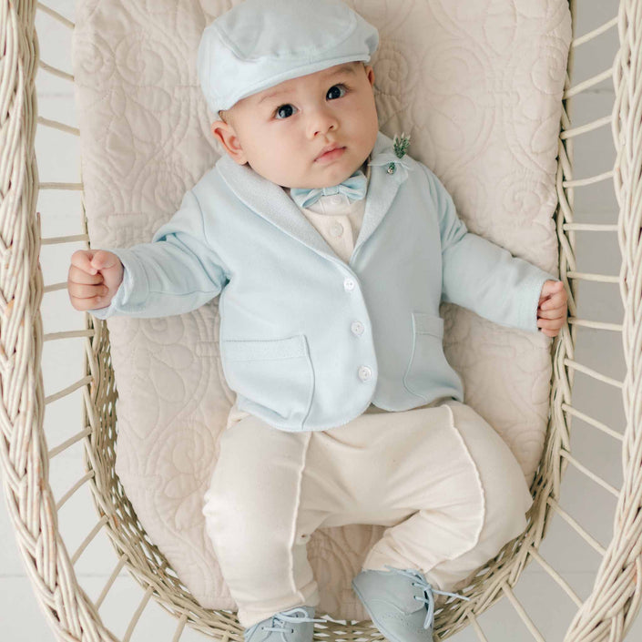 A baby dressed in an Ezra Powder Blue 4-Piece Suit, featuring a light blue jacket made from French Terry Cotton, white pants, and a matching light blue cap is sitting in a wicker chair. The baby has a neutral expression and is holding onto the sides of the chair. The background is light and neutral.