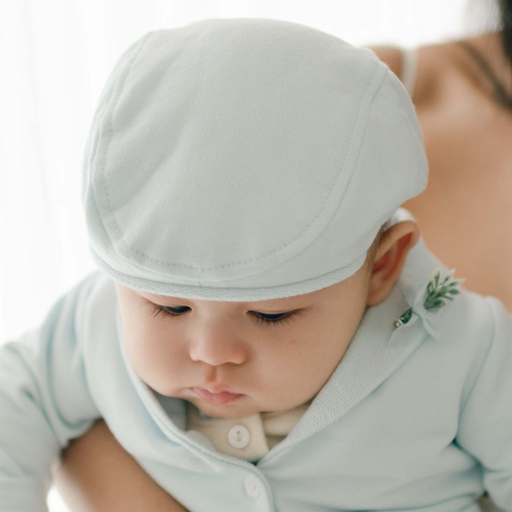 A baby wearing a light blue outfit and an Ezra Powder Blue Newsboy Cap is being held by an adult. The baby is looking downward with a calm expression. The background is softly blurred, putting focus on the baby, enhancing the serene christening look.
