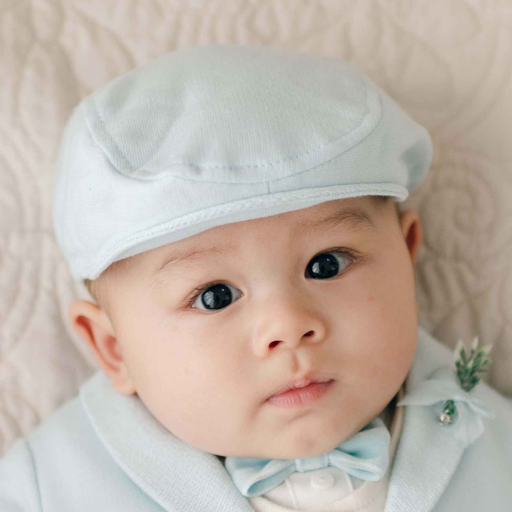 An infant wearing an Ezra Powder Blue Newsboy Cap and matching light blue outfit, including a bow tie, looks toward the camera. The baby has a neutral expression and is positioned against a fabric background.