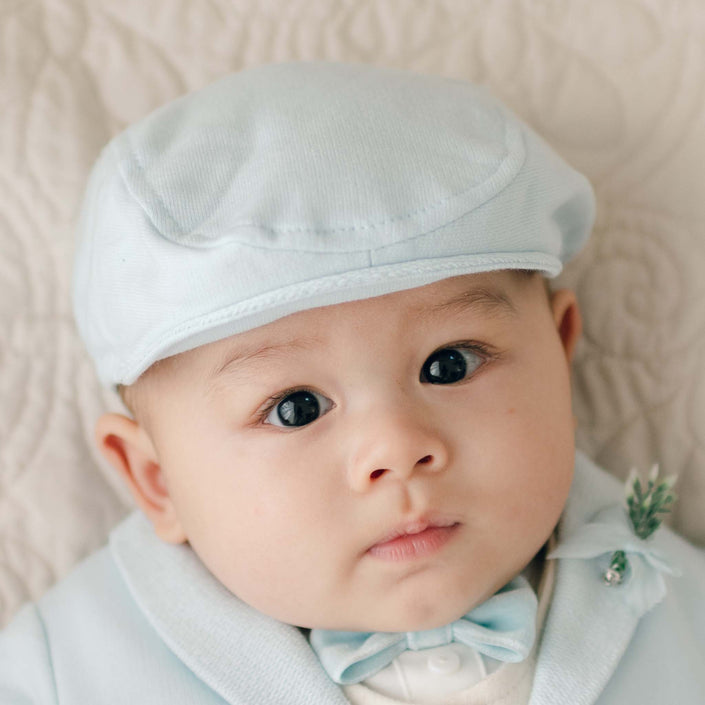 An infant wearing an Ezra Powder Blue Newsboy Cap and matching light blue outfit, including a bow tie, looks toward the camera. The baby has a neutral expression and is positioned against a fabric background.