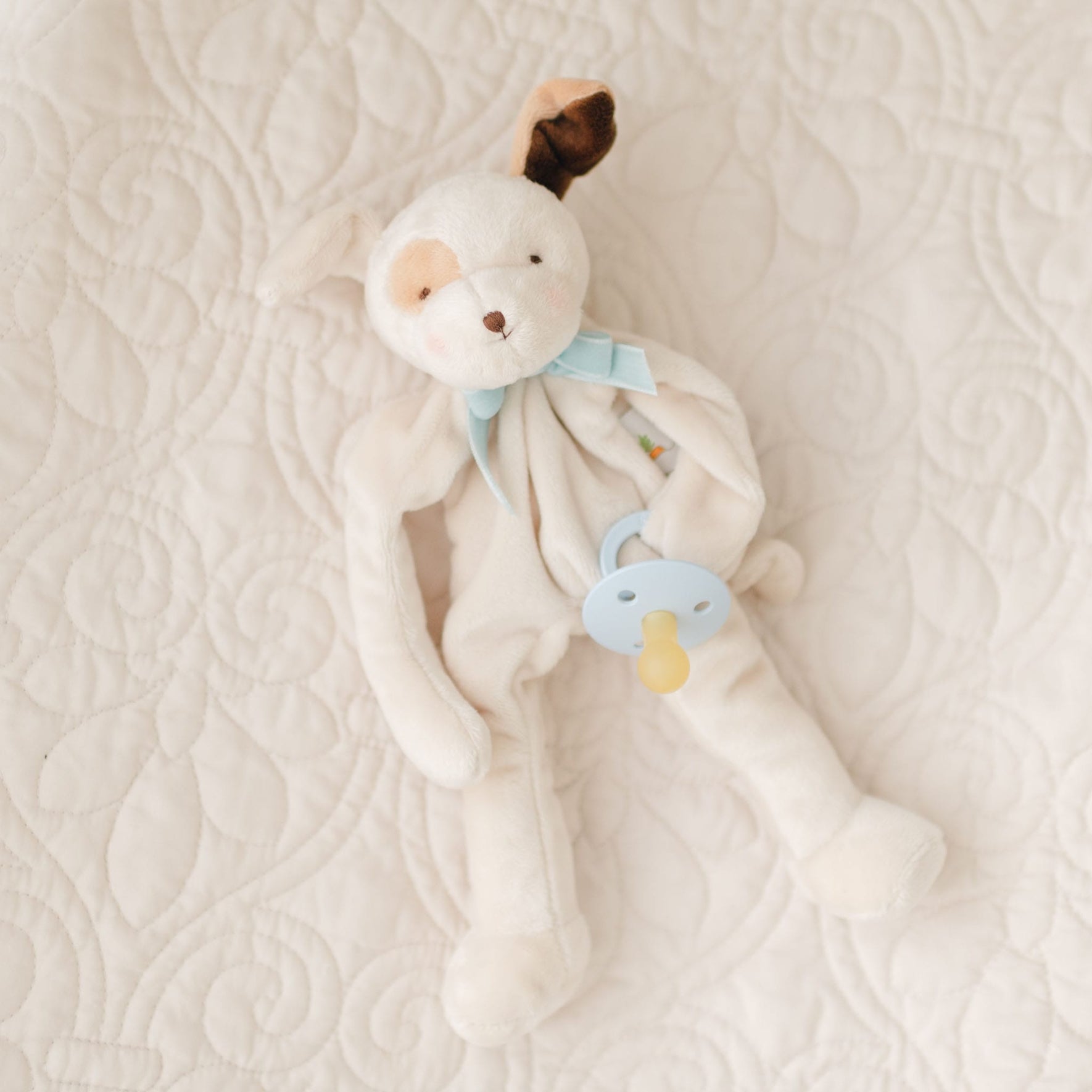A soft, cream-colored plush toy resembling a dog lies on a quilted white bedspread. The toy has floppy ears, one brown and one cream, and wears a blue ribbon around its neck. Designed as a delightful baby gift, the Ezra Powder Blue Silly Puppy Buddy | Pacifier Holder holds a blue pacifier with a yellow tip in one paw.