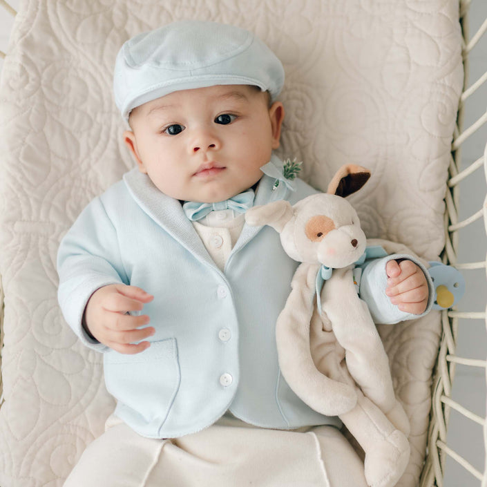 A baby dressed in a light blue suit jacket, matching hat, and bowtie is lying on a quilted surface. The baby is looking up while holding an Ezra Powder Blue Silly Puppy Buddy | Pacifier Holder.