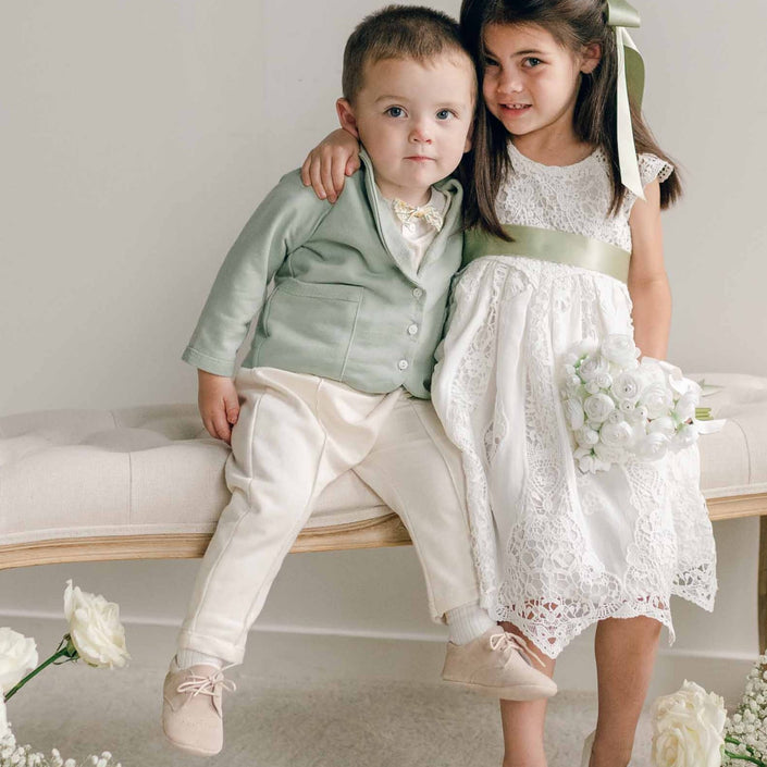 A young boy wearing the Ezra Sage 4-Piece Suit sits on a tufted bench beside a girl in a lace dress with a sage sash, surrounded by white roses.
