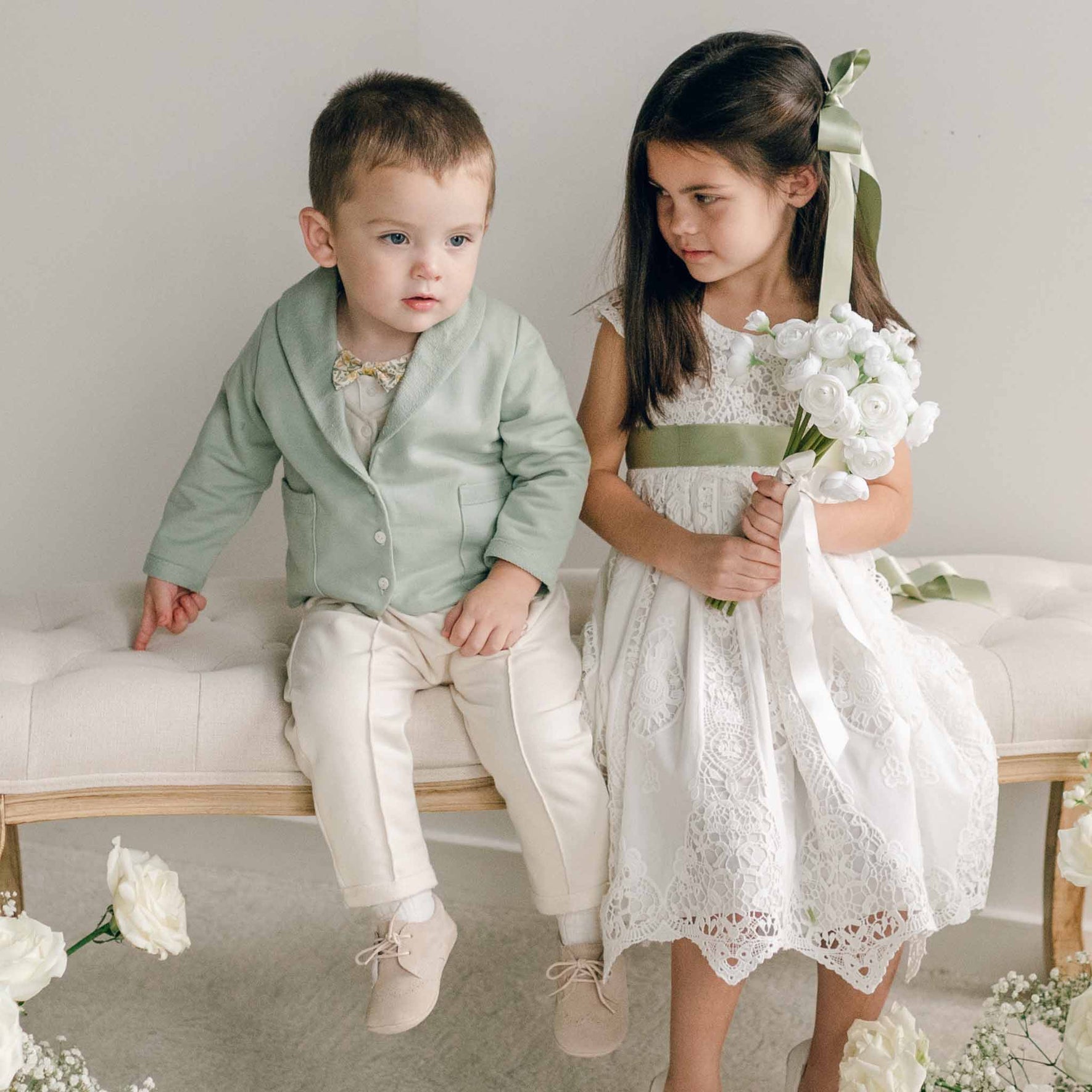 A young boy in the Ezra Sage 4-Piece Suit and a girl in a lace dress with a sage sash sit together on a bench, with the girl holding a white floral bouquet.