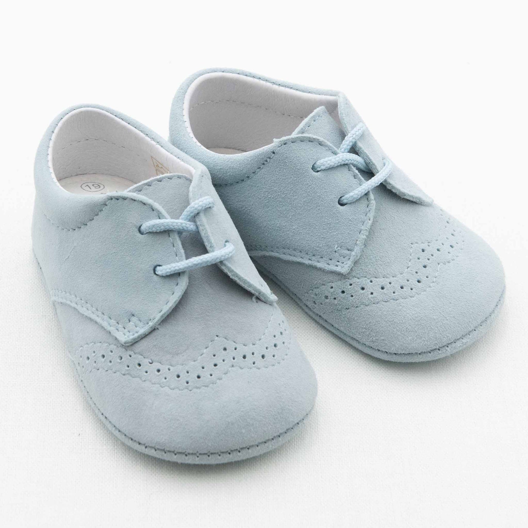 A pair of light blue Ezra Suede Shoes is laid on a plain white background. The handmade suede shoes feature laces and decorative perforations on the toe and side areas, with a soft, suede-like texture. The size label inside reads "18.