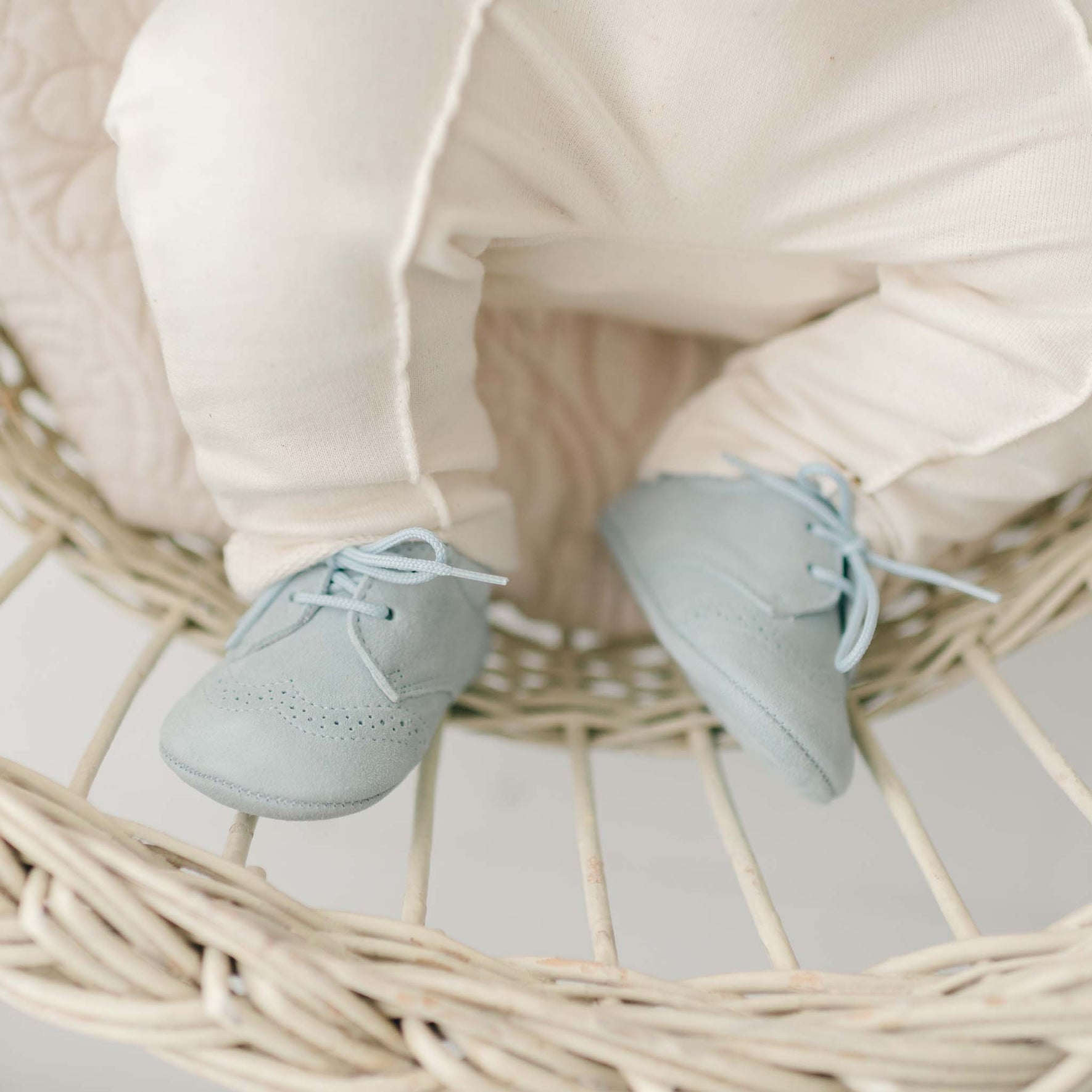 An infant wearing cream-colored pants and light blue Ezra Suede Shoes sits in a wicker basket. Only the lower half of the baby's body is visible, with the shoes' laces neatly tied.