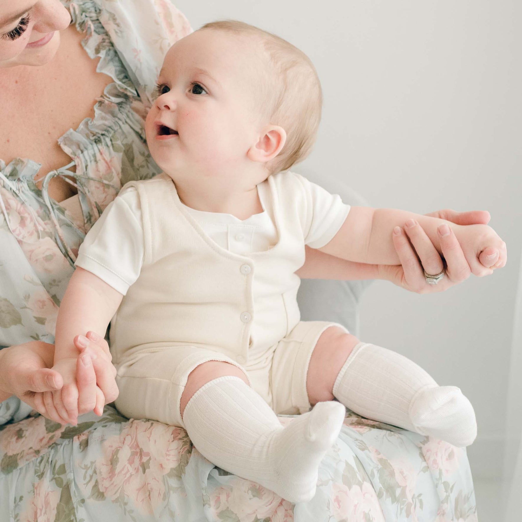 A baby in the Ezra Tan 4-Piece Suit sits on a mother's lap, smiling while holding her hands, wearing an ivory vest and shorts.