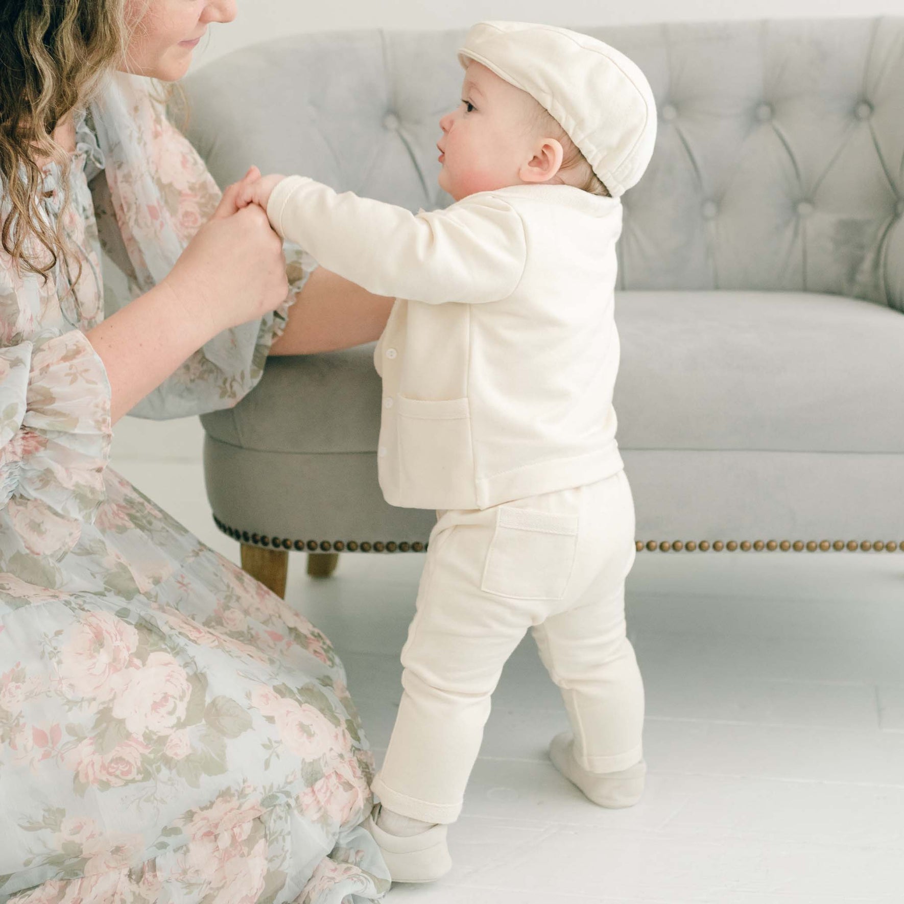 A baby wearing the Ezra Tan 4-Piece Suit stands with support, showing the back of the soft ivory outfit with a buttoned vest and matching pants.