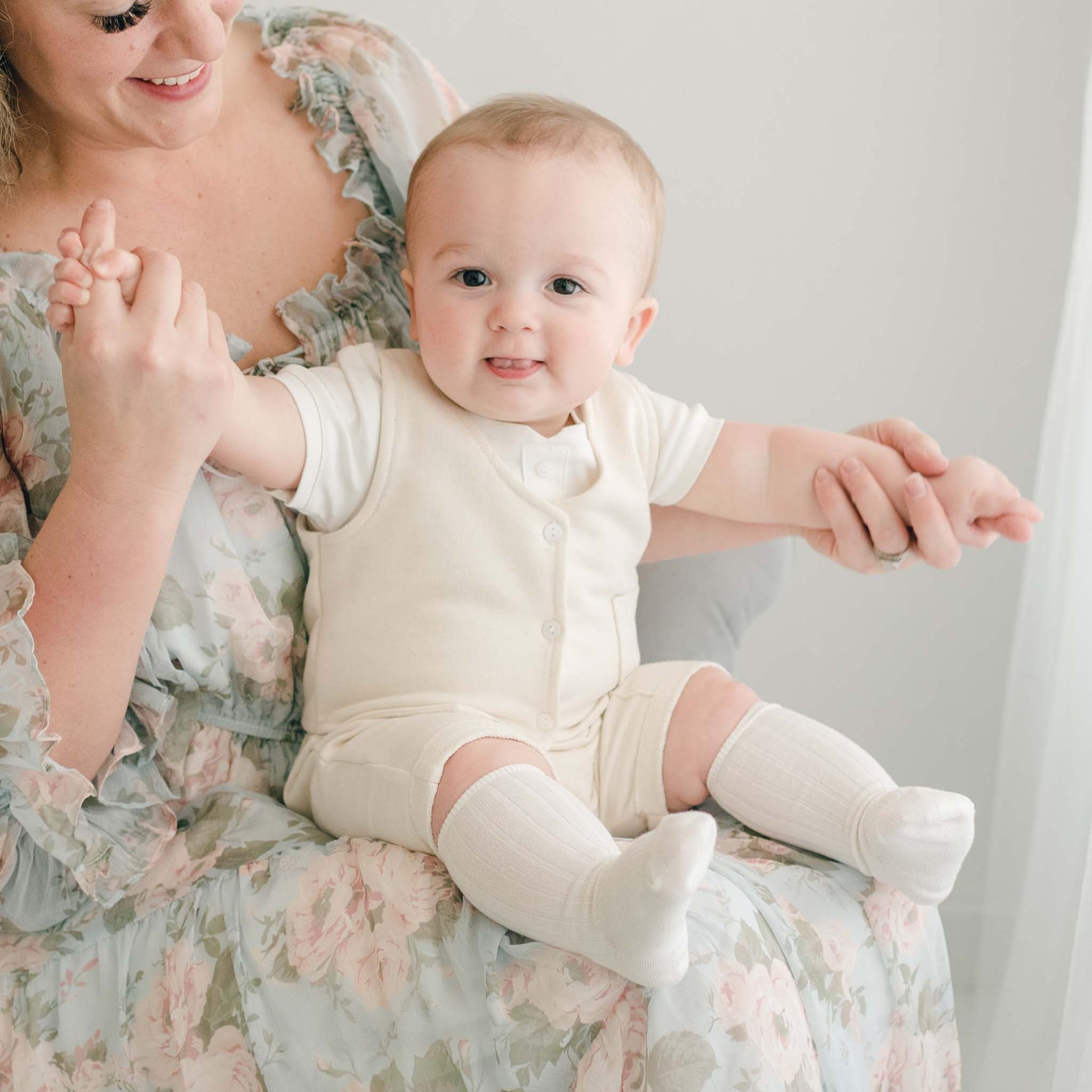 A baby in the Ezra Tan 4-Piece Suit sits on a mother's lap, holding her hands, wearing a soft ivory outfit with a buttoned vest.