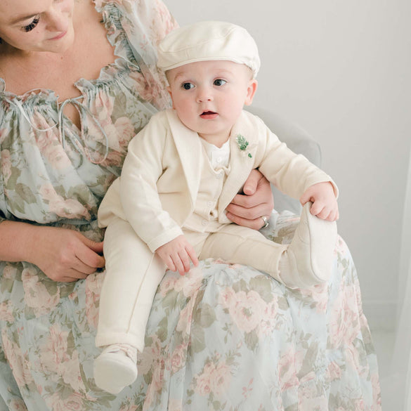 A baby in the Ezra Tan 4-Piece Suit sits on a mother's lap, dressed in an ivory ensemble with a matching cap and shoes.