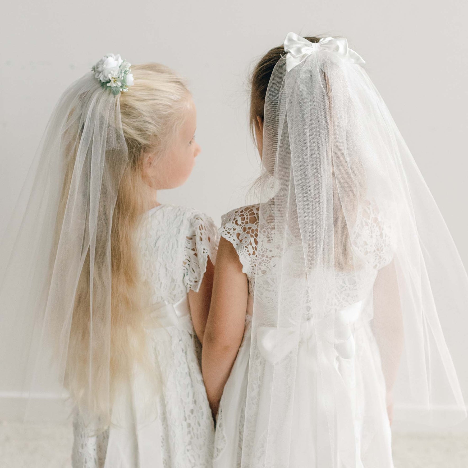 Two young girls sit side by side, wearing the First Communion Veil with their white lace dresses.