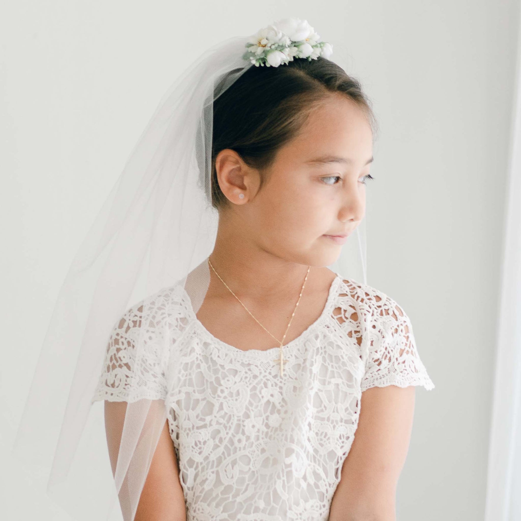 A girl wears the First Communion Veil with a floral headpiece, paired with a white lace dress.