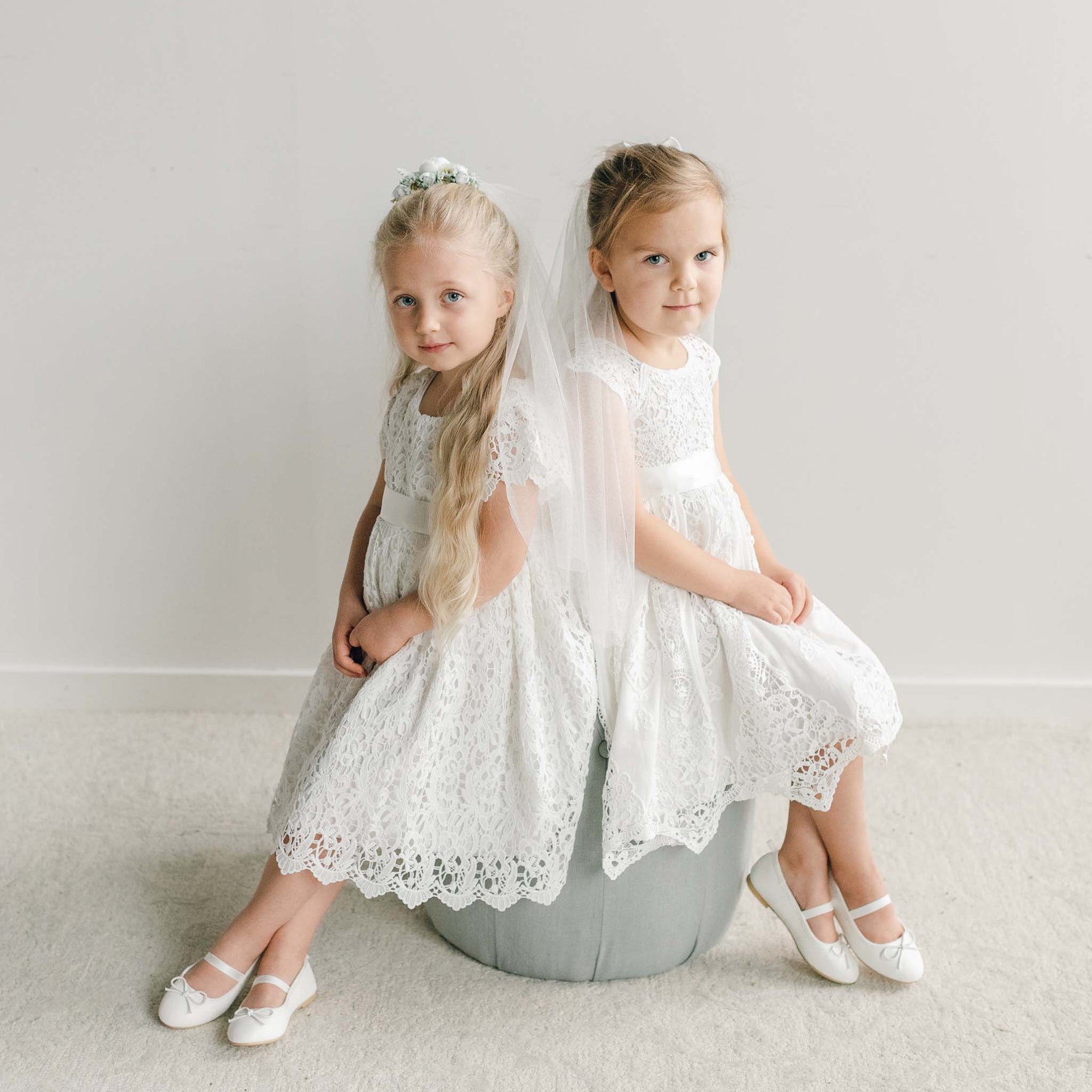 Two girls in matching lace dresses and satin sashes sit together, one wearing a white floral veil and the other a bow with a flowing veil.