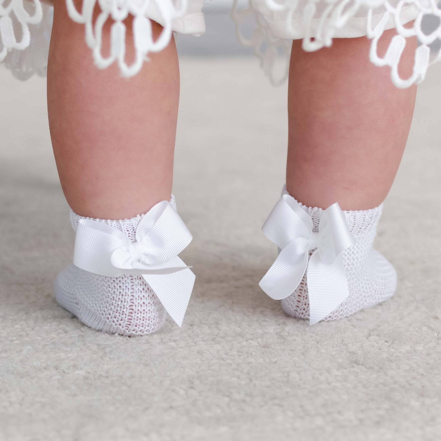 A close-up of a baby's legs wearing Garter Stitch Socks with Bow, featuring large grosgrain ribbon bows on the back. The baby stands on a light-colored carpet, with part of a white lace dress visible.