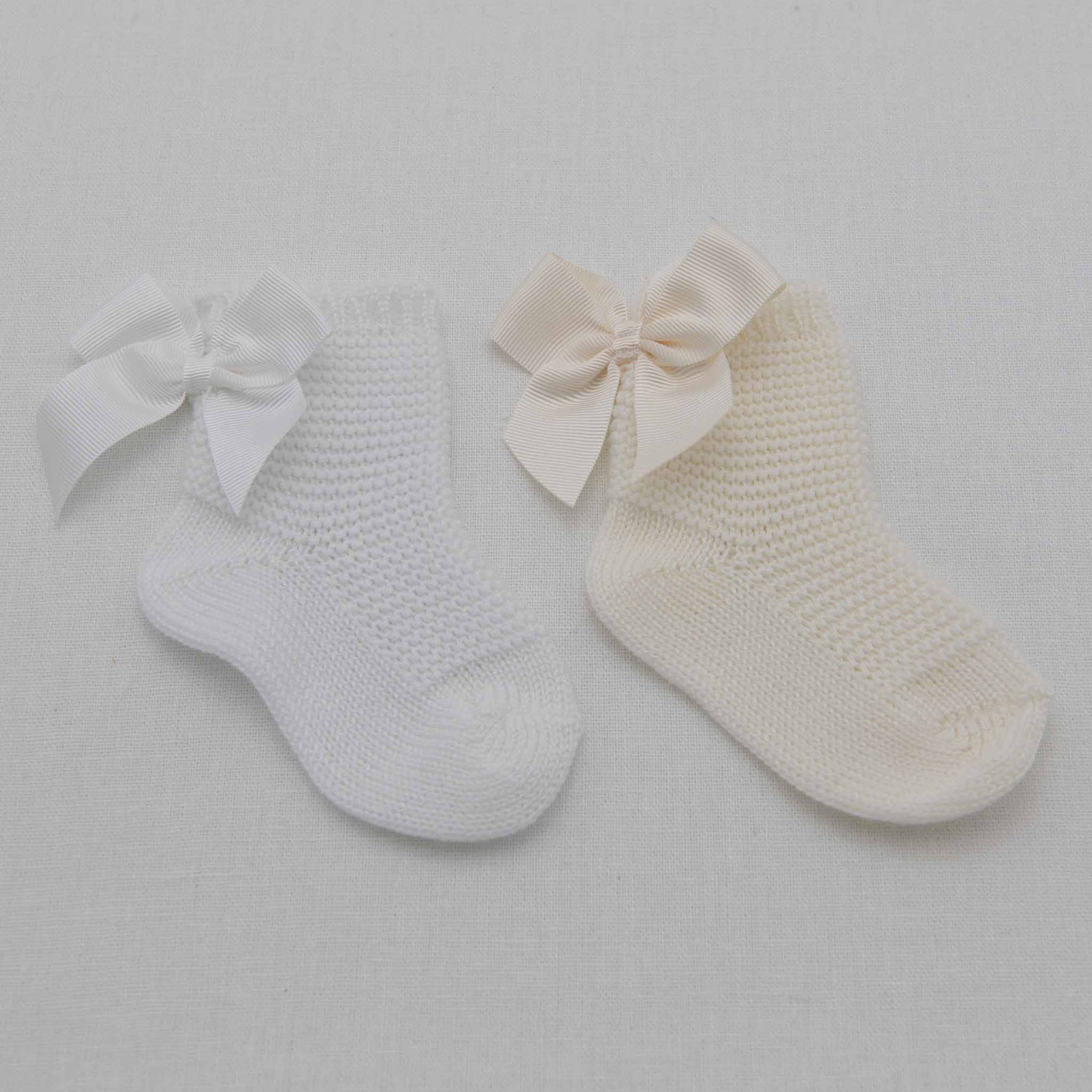 The image shows two Garter Stitch Socks with Bow against a white background. The sock on the left is white, 100% cotton, with a matching grosgrain ribbon bow, while the sock on the right is cream with a cream bow.