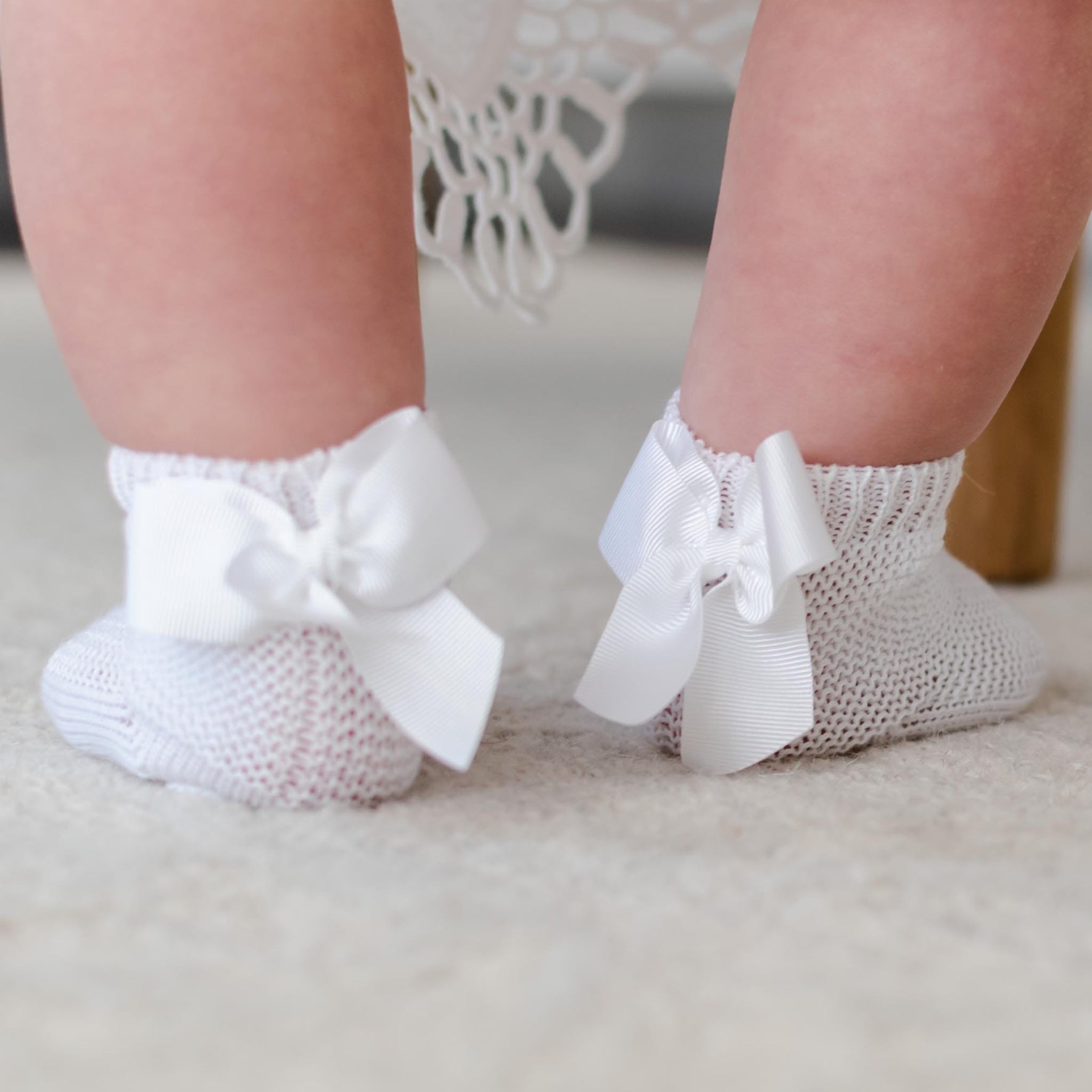 Close-up of a baby's legs wearing Garter Stitch Socks with Bow, made from 100% cotton, featuring large grosgrain ribbon bows. The socks are standing on a soft, light-colored carpet against a backdrop of draped fabric.