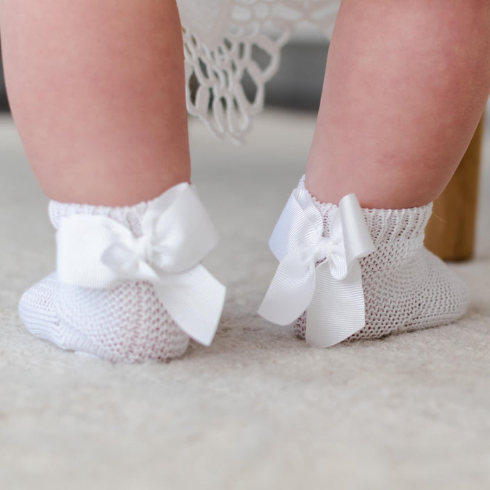 Close-up of a baby's legs wearing Garter Stitch Socks with Bow, made from 100% cotton, featuring large grosgrain ribbon bows. The socks are standing on a soft, light-colored carpet against a backdrop of draped fabric.