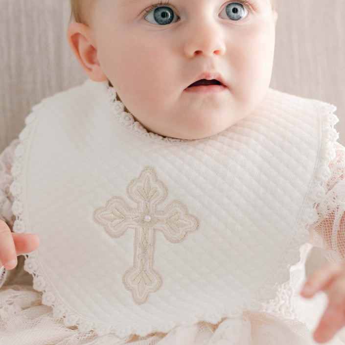 A baby with light hair and blue eyes is wearing a white lace dress featuring an embroidered cross pattern on the front. Enhanced with pearl-style bead details, the ensemble adds elegance as the baby gazes slightly to the side against a soft, neutral background. The outfit matches perfectly with the Girls Cross Bib for a cohesive look.