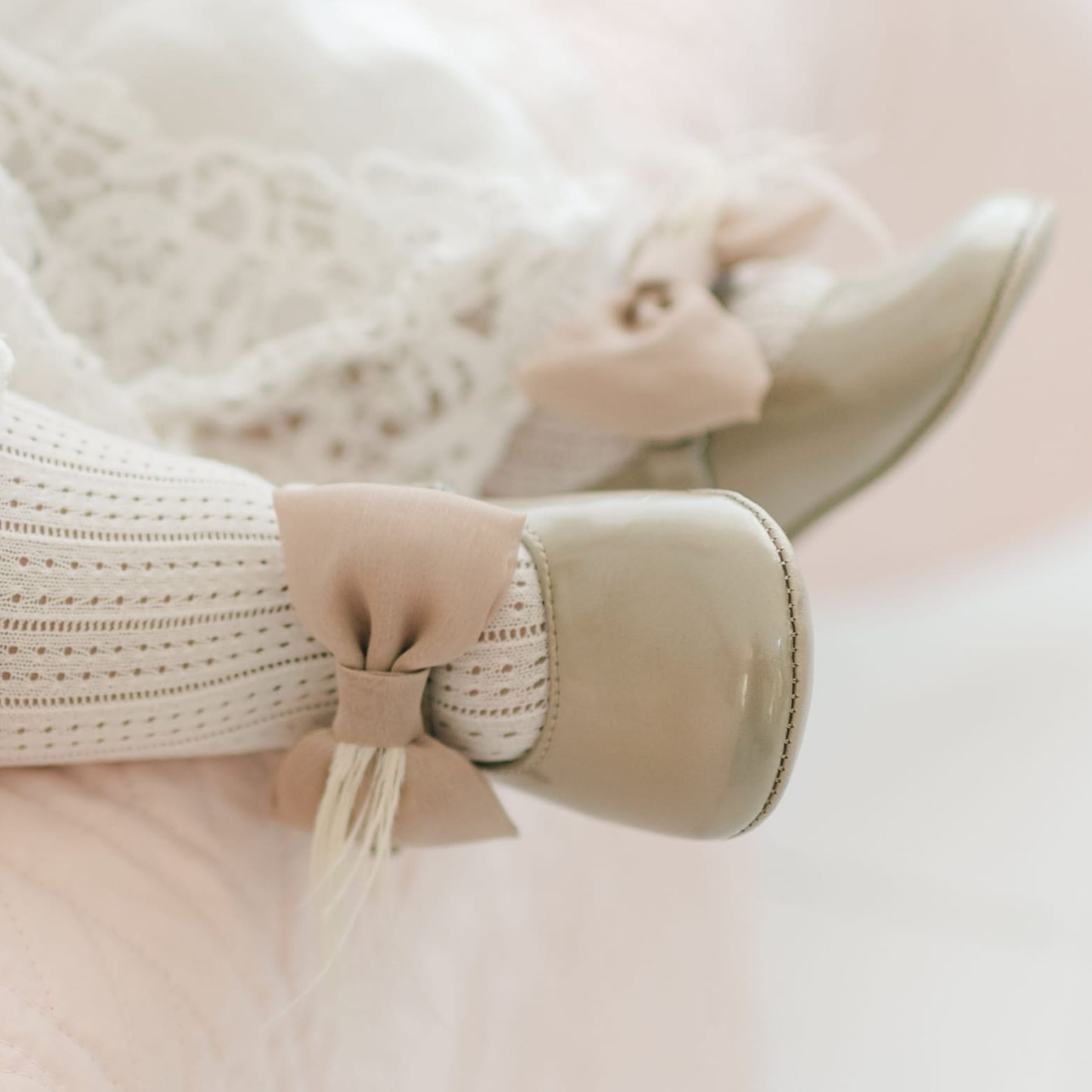Close-up a baby's feet wearing the Gold Patent Leather Mary Janes. They are made with gold patent leather with a soft velcro strap that features a champagne chiffon bow and feather. The background shows a light pink quilt.