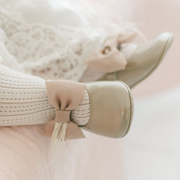 Close-up a baby's feet wearing the Gold Patent Leather Mary Janes. They are made with gold patent leather with a soft velcro strap that features a champagne chiffon bow and feather. The background shows a light pink quilt.