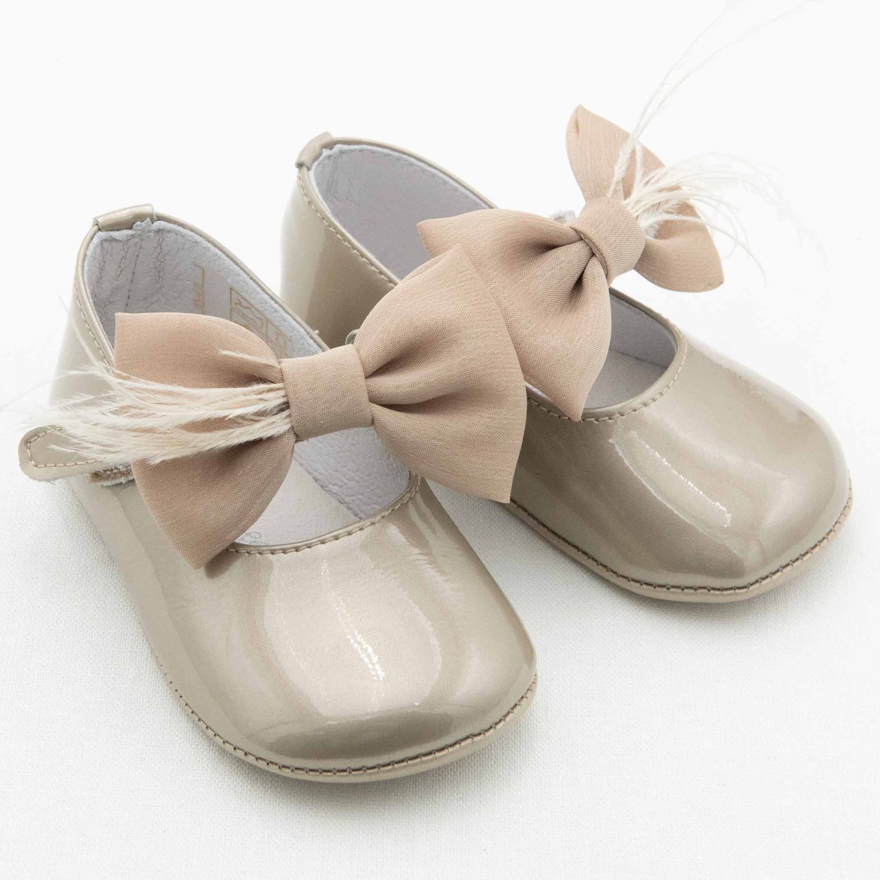 A pair of small Gold Patent Leather Mary Janes. The shoes are made with gold patent leather with a soft velcro strap that features a coordinating champagne chiffon bow and feather. The background is white.