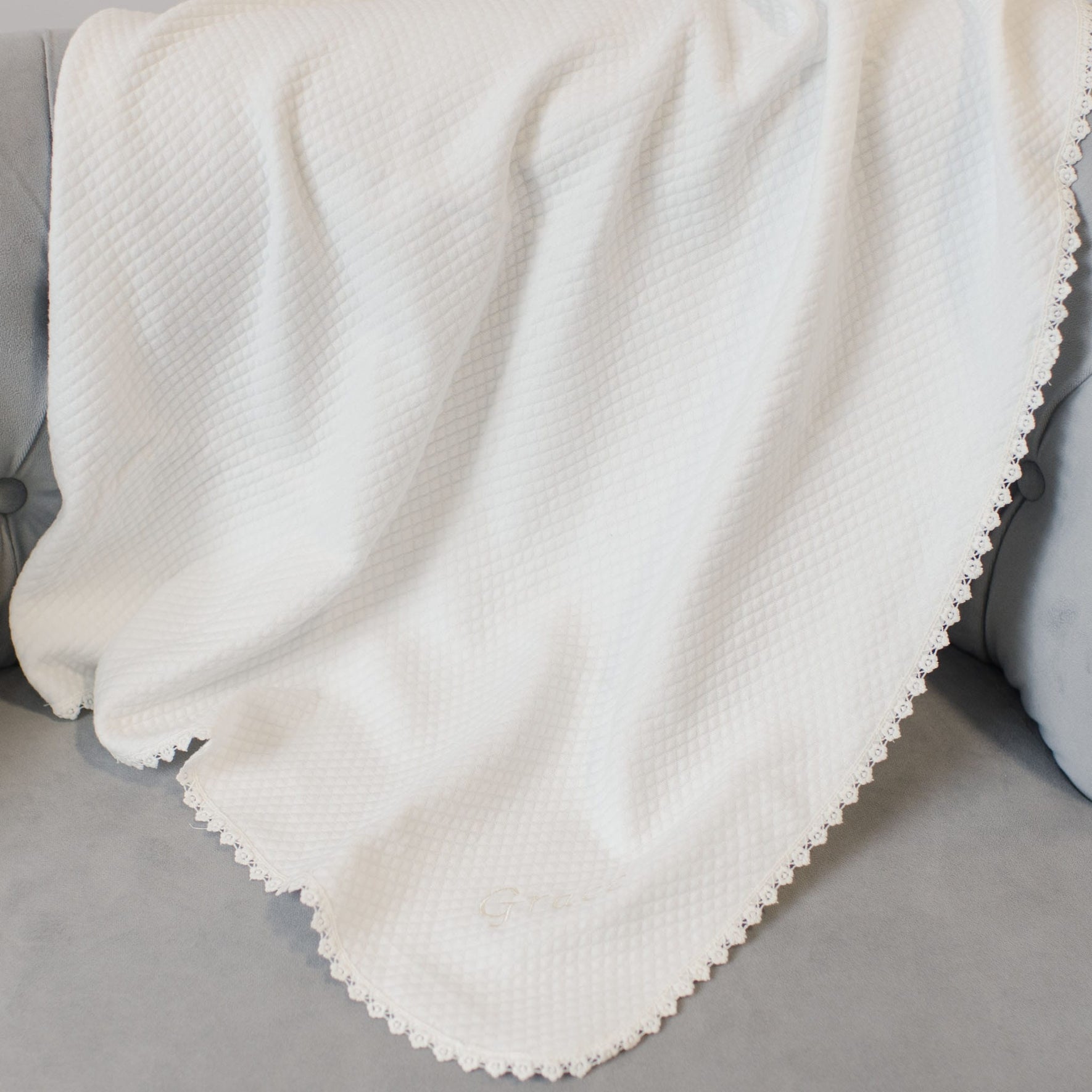 The Grace Accessory Bundle, featuring a personalized quilted blanket with a white waffle-pattern and lace trim, is elegantly draped over a gray sofa.