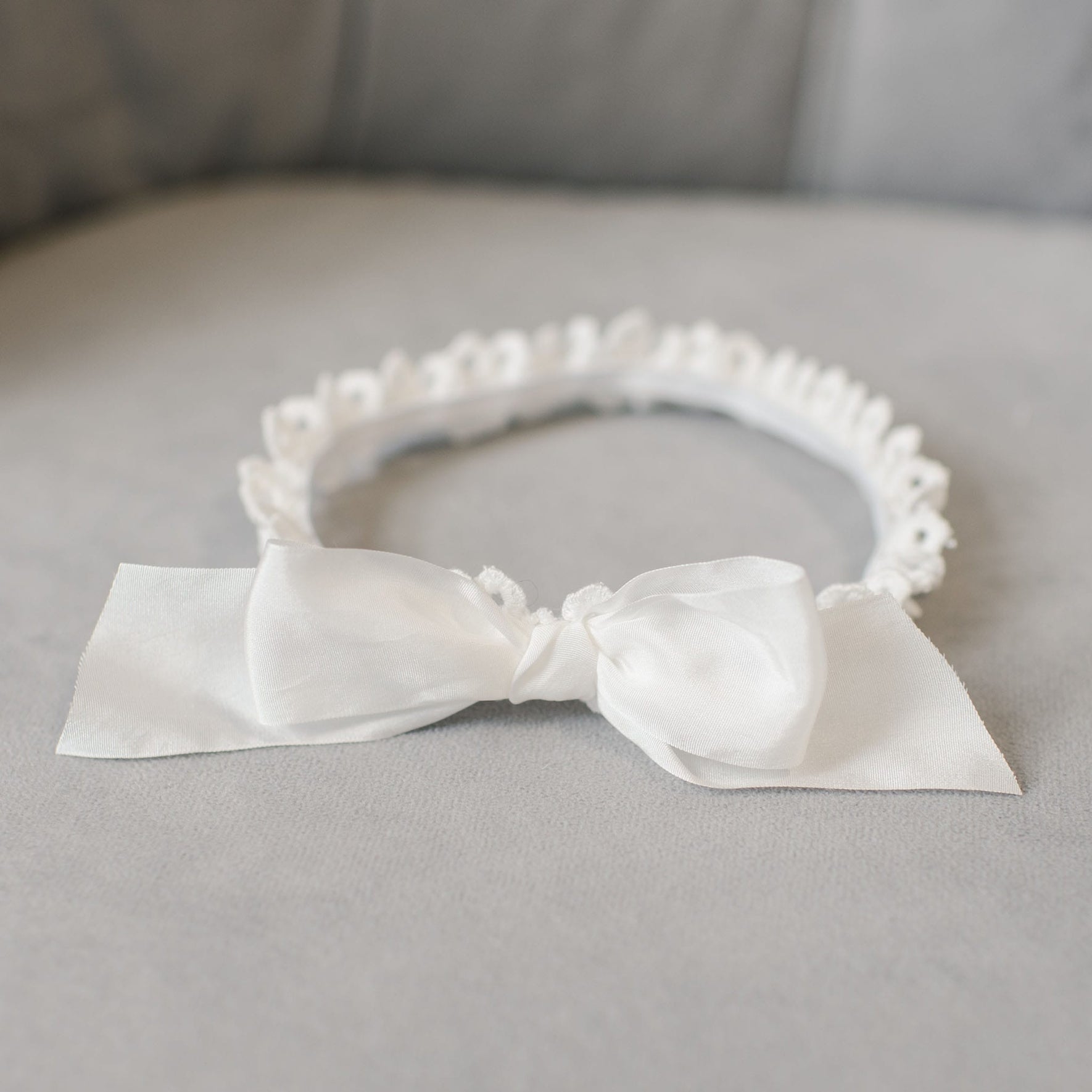A white headband featuring a large bow rests on a light gray fabric surface, enhancing the intricate details of the Grace Christening gown beautifully. The headband is adorned with small decorative elements along its edge, making it an ideal addition to the Grace Accessory Bundle - Save 15% for a baptism accessory set.