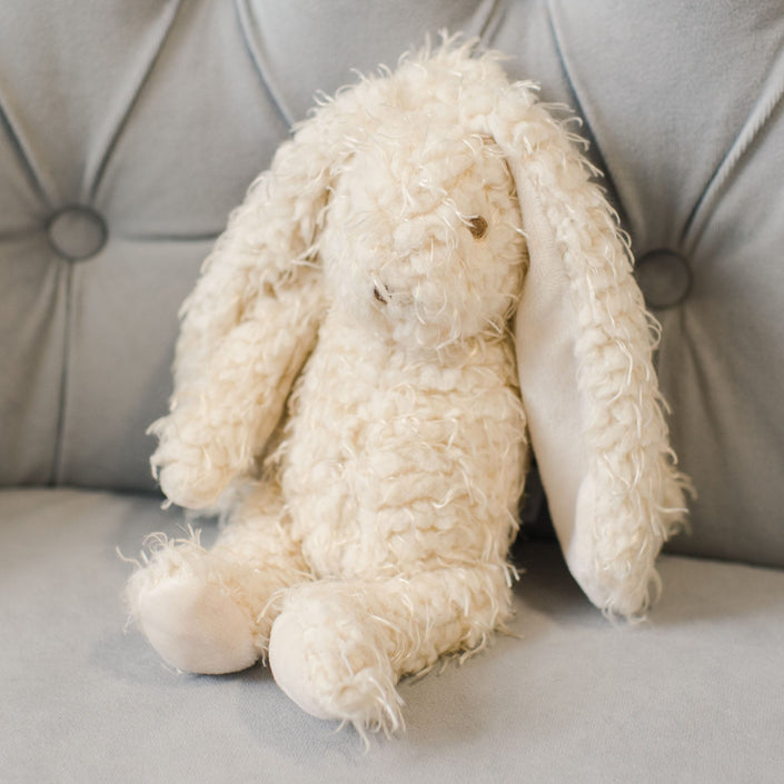 The Grace Bunny, with its cream-colored textured fur-like appearance and irresistibly soft, fluffy fur, sits gracefully on a light gray cushioned surface. Its long ears make it an ideal baby gift that invites endless cuddles.