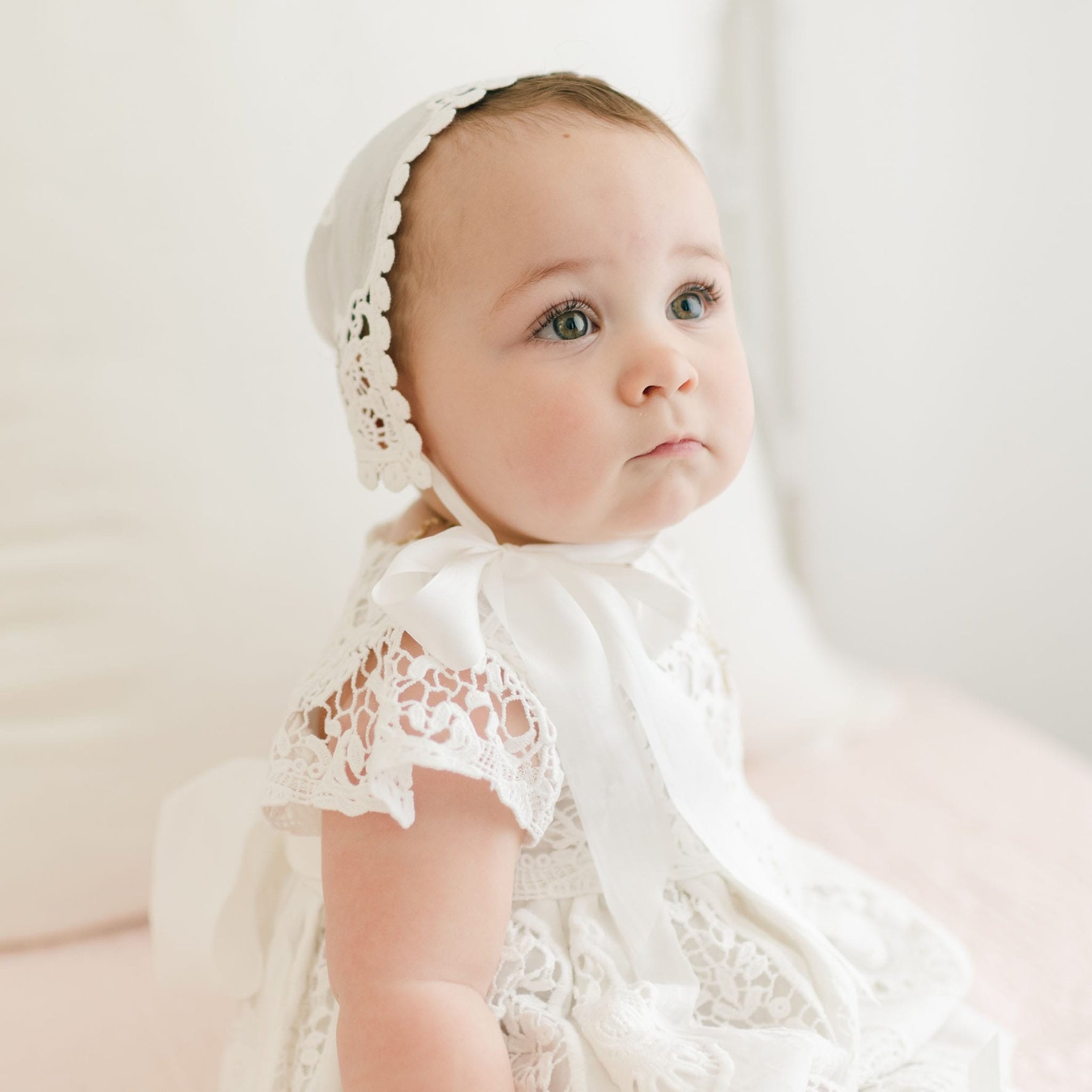 A light-skinned baby with light brown hair is seated, wearing a white lace bonnet and the Grace Christening Dress. The dress showcases a delicate lace bodice, and the baby gazes slightly upward with a neutral expression. The softly blurred background features white and pastel tones.