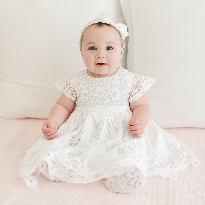 A baby sits on a light pink surface adorned with the Grace Christening Dress, featuring a white crochet design and a lace bodice, complemented by a matching headband with a bow. The baby has light skin and blue eyes and is looking forward with a neutral expression, surrounded by white cushions in the background.