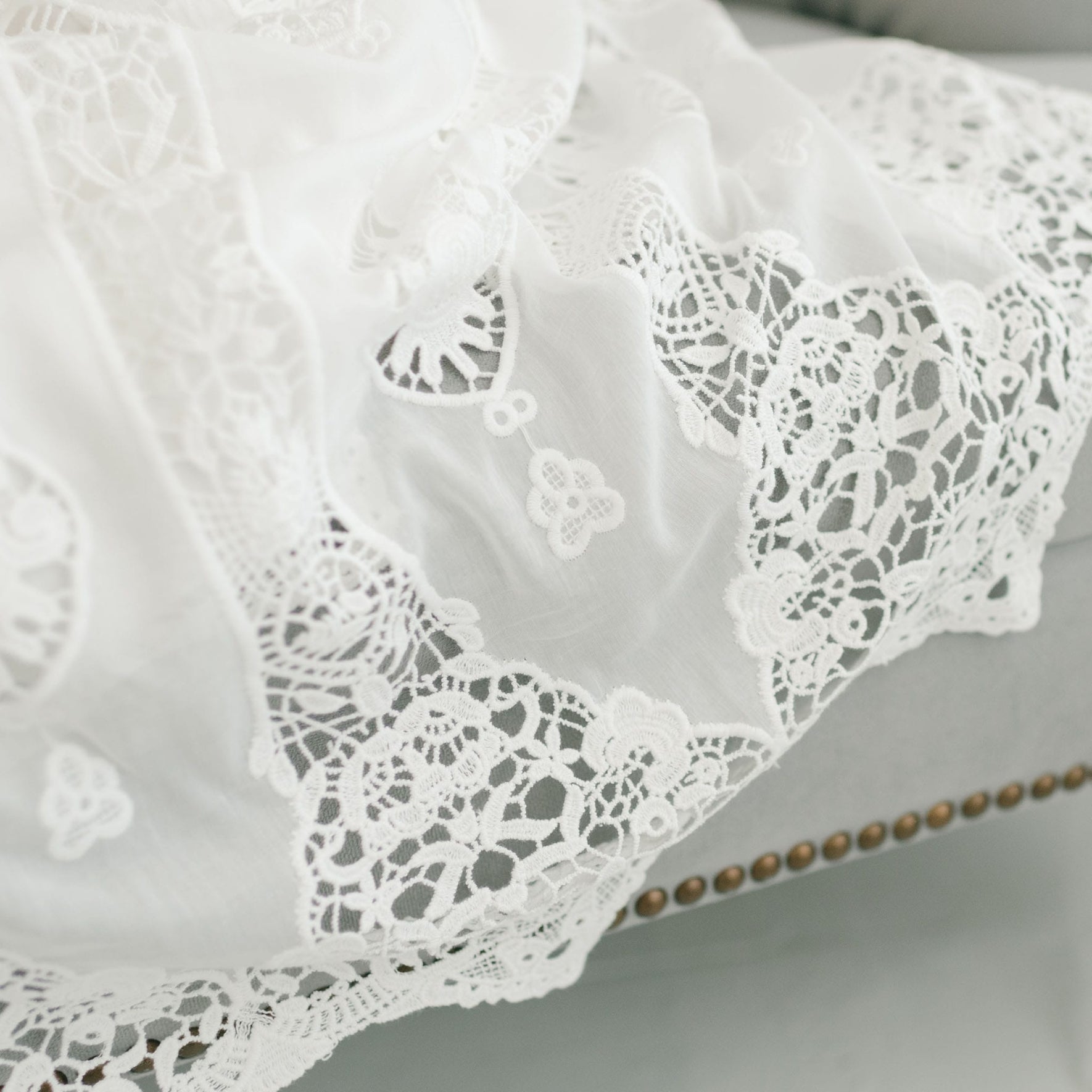 Close-up of Grace Christening Gown & Bonnet draped over a light gray piece of furniture. The lace, reminiscent of a Christening Bonnet, features intricate floral patterns and delicate detailing, adding a touch of elegance to the scene. The edge of the fabric appears to be finished with scalloped designs.