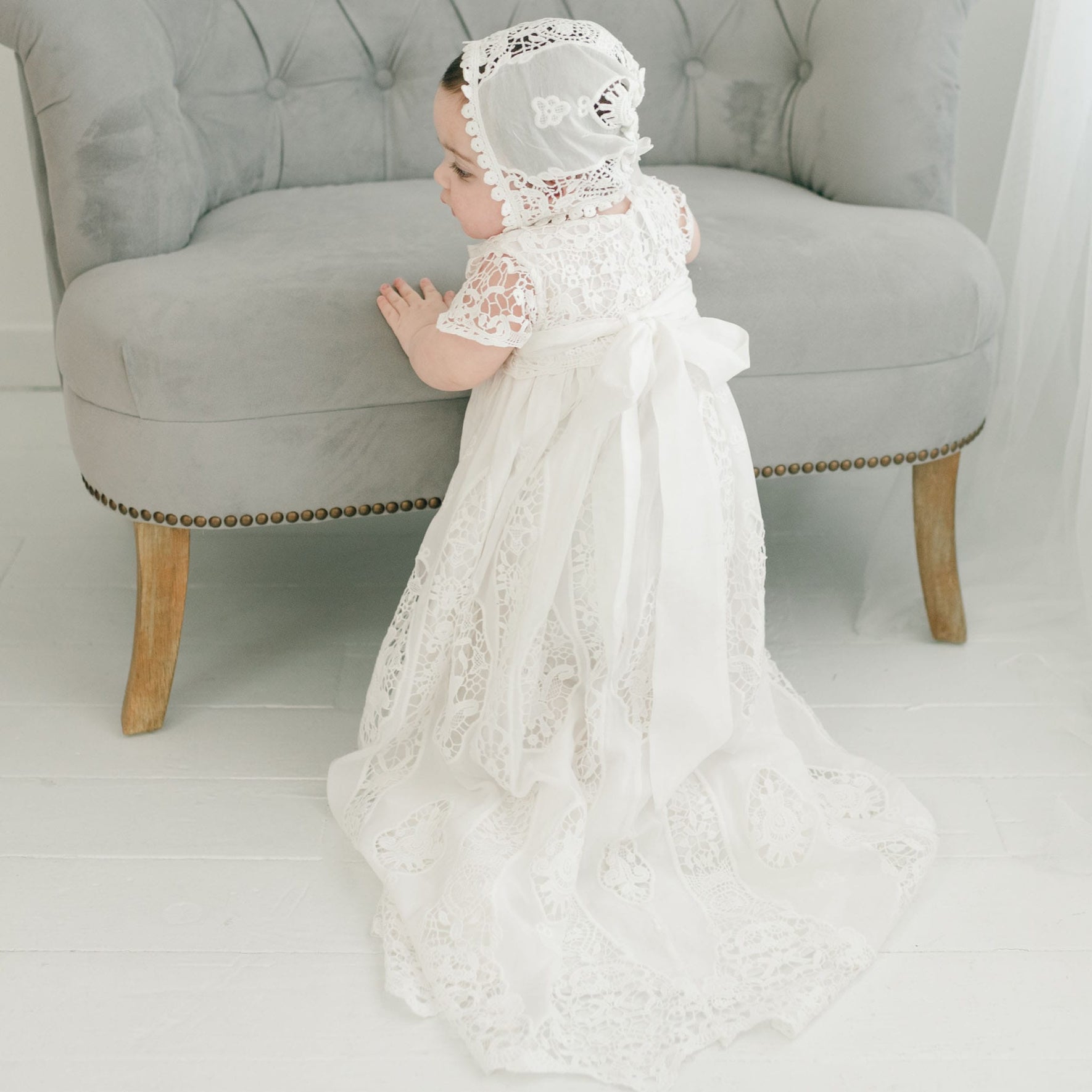 A young child stands facing a gray upholstered chair, dressed in the exquisite Grace Christening Gown & Bonnet. The detailed lace gown features a long train and is complemented beautifully by a matching bonnet adorned with delicate patterns. A silk sash adds an elegant touch to the outfit. The scene unfolds against a backdrop of a white floor and sheer curtains.