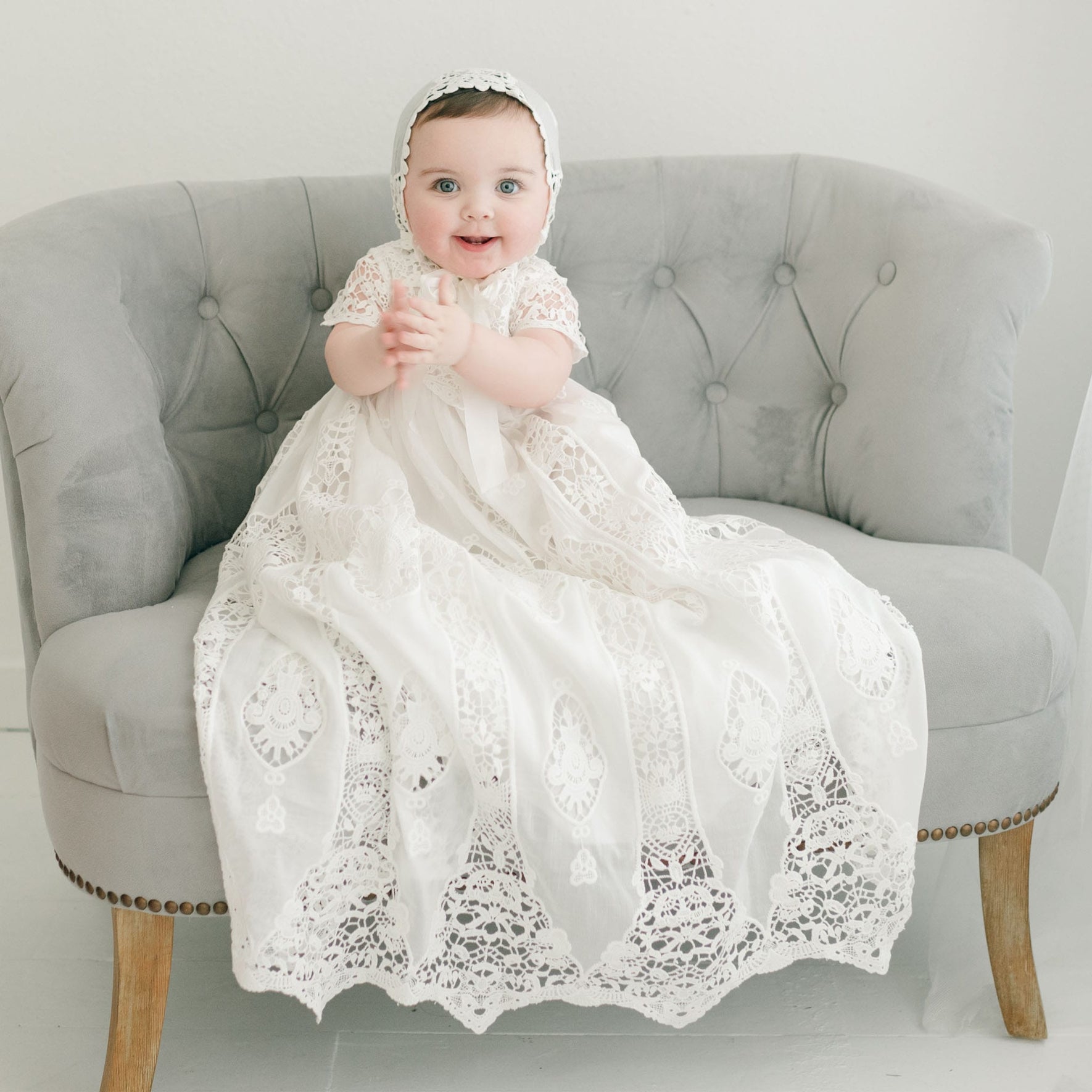 A baby is sitting on a tufted, light grey couch, wearing the Grace Christening Gown & Bonnet. The baby has a joyful expression, clapping hands against a plain white indoor background.