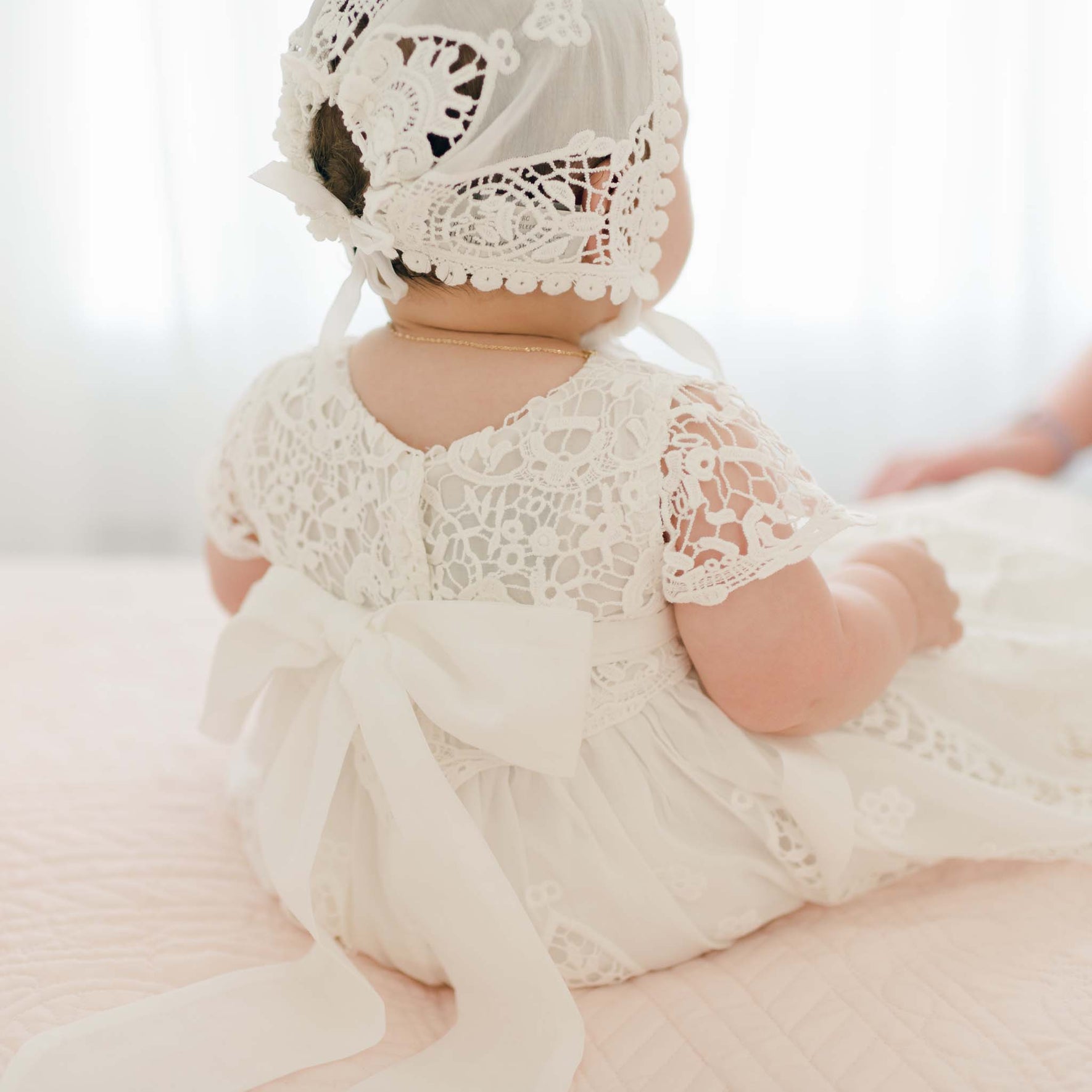 A baby dressed in the Grace Christening Gown & Bonnet sits on a light-colored quilted surface. The back of the elegant lace gown is adorned with a large white bow, though the baby's face is not visible.