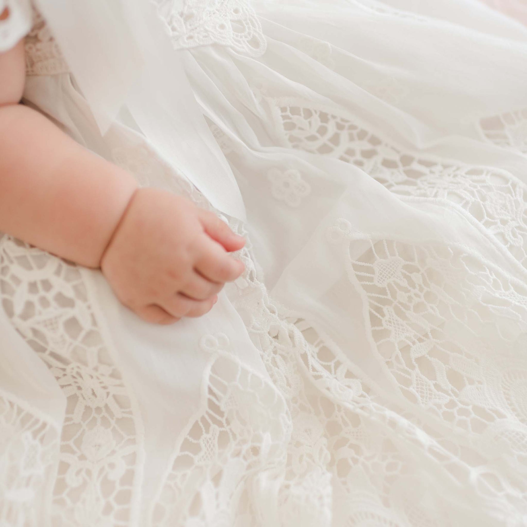Close-up of a baby's hand resting on the Grace Christening Gown & Bonnet, showcasing its intricate floral patterns and layers of fine lace fabric. A silk sash adorns the waist, adding an extra touch of elegance. The baby’s arm is visible, showing a soft, chubby appearance.