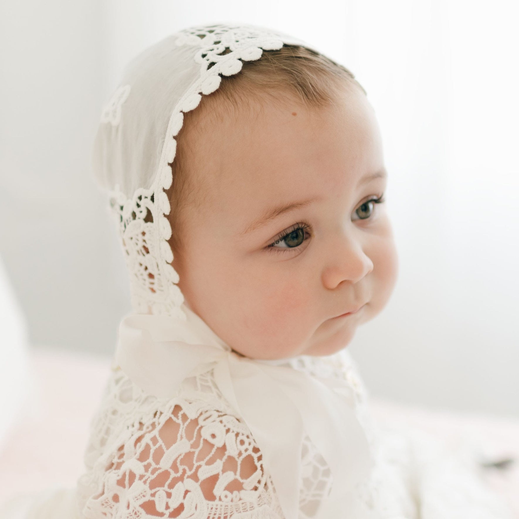 A baby is wearing the Grace Christening Gown & Bonnet, featuring intricate white lace and a delicate silk sash. The baby gazes slightly to the side with a neutral expression, while the softly lit background remains out of focus.