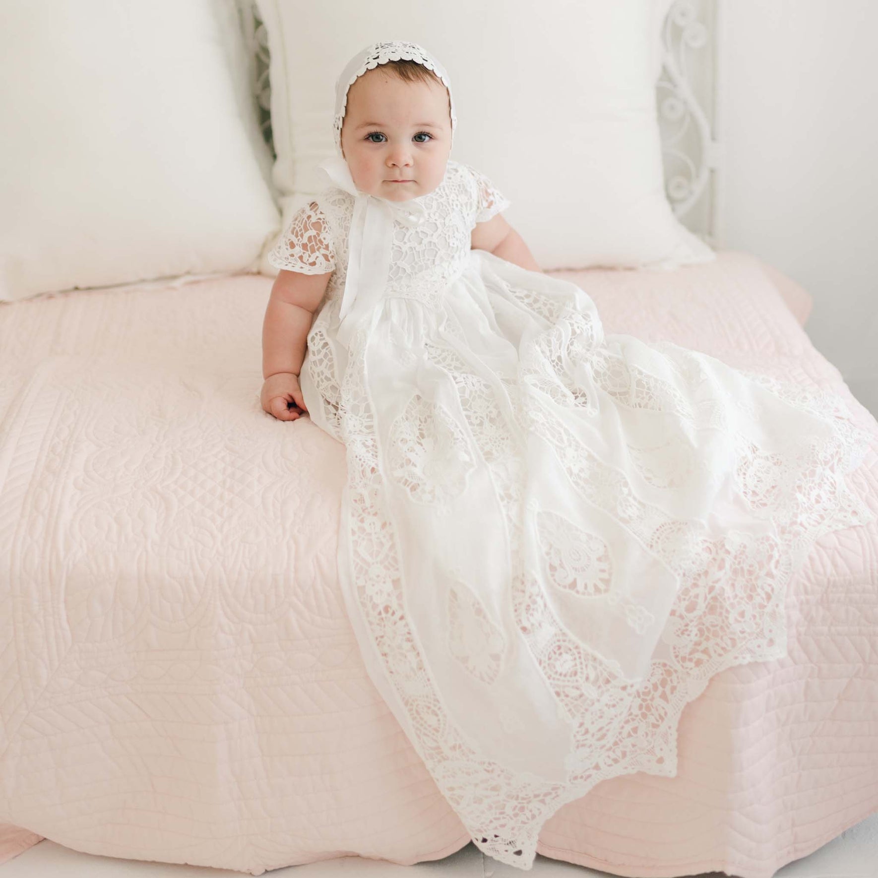A baby wearing the Grace Christening Gown & Bonnet sits on a blush pink quilted blanket. The baby has a neutral expression and is positioned against a backdrop of white pillows and a white headboard.
