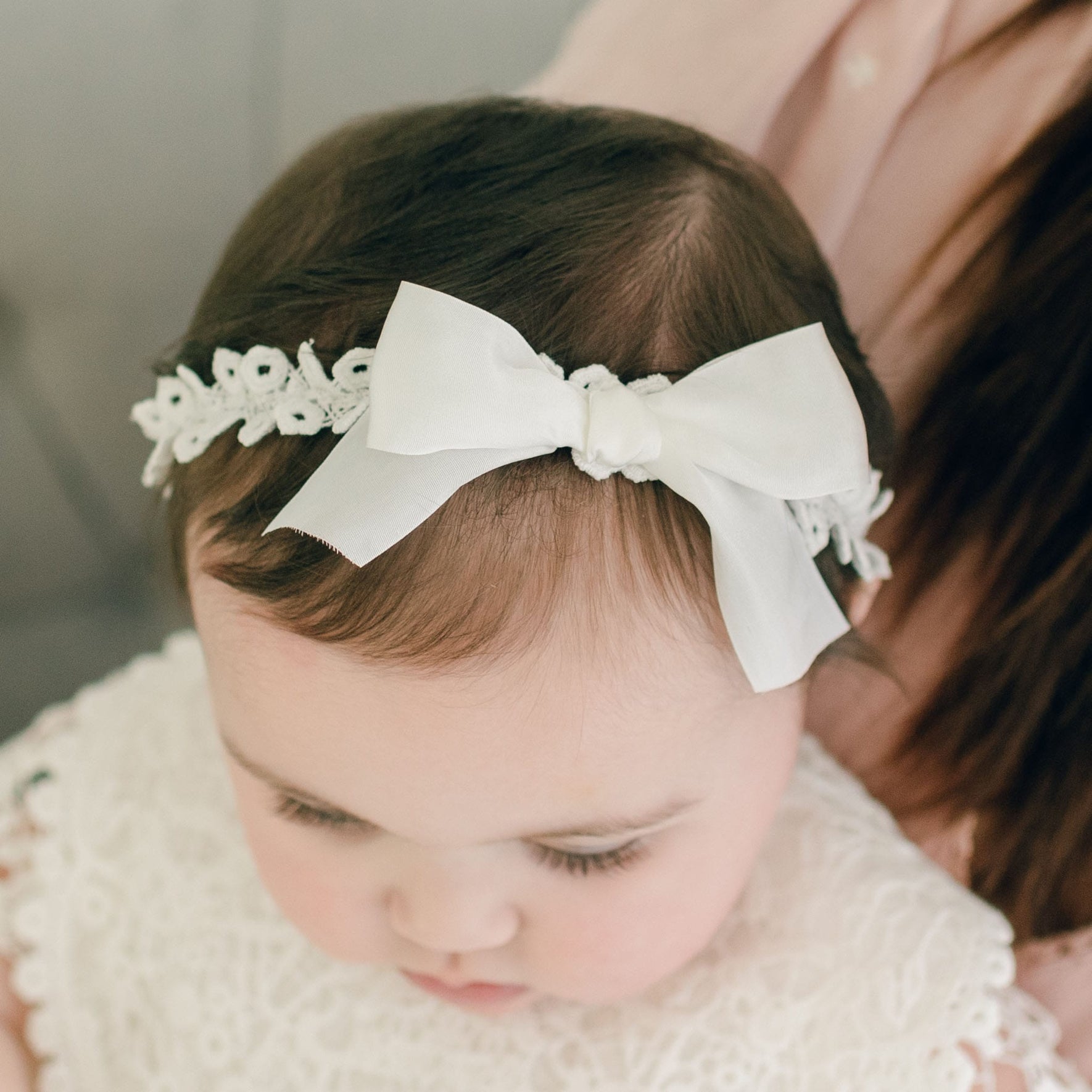 A baby is wearing the Grace Christening Headband, adorned with a silk ribbon bow and lace. Dressed in a white outfit, the baby is looking down while an adult, partially visible, holds them.