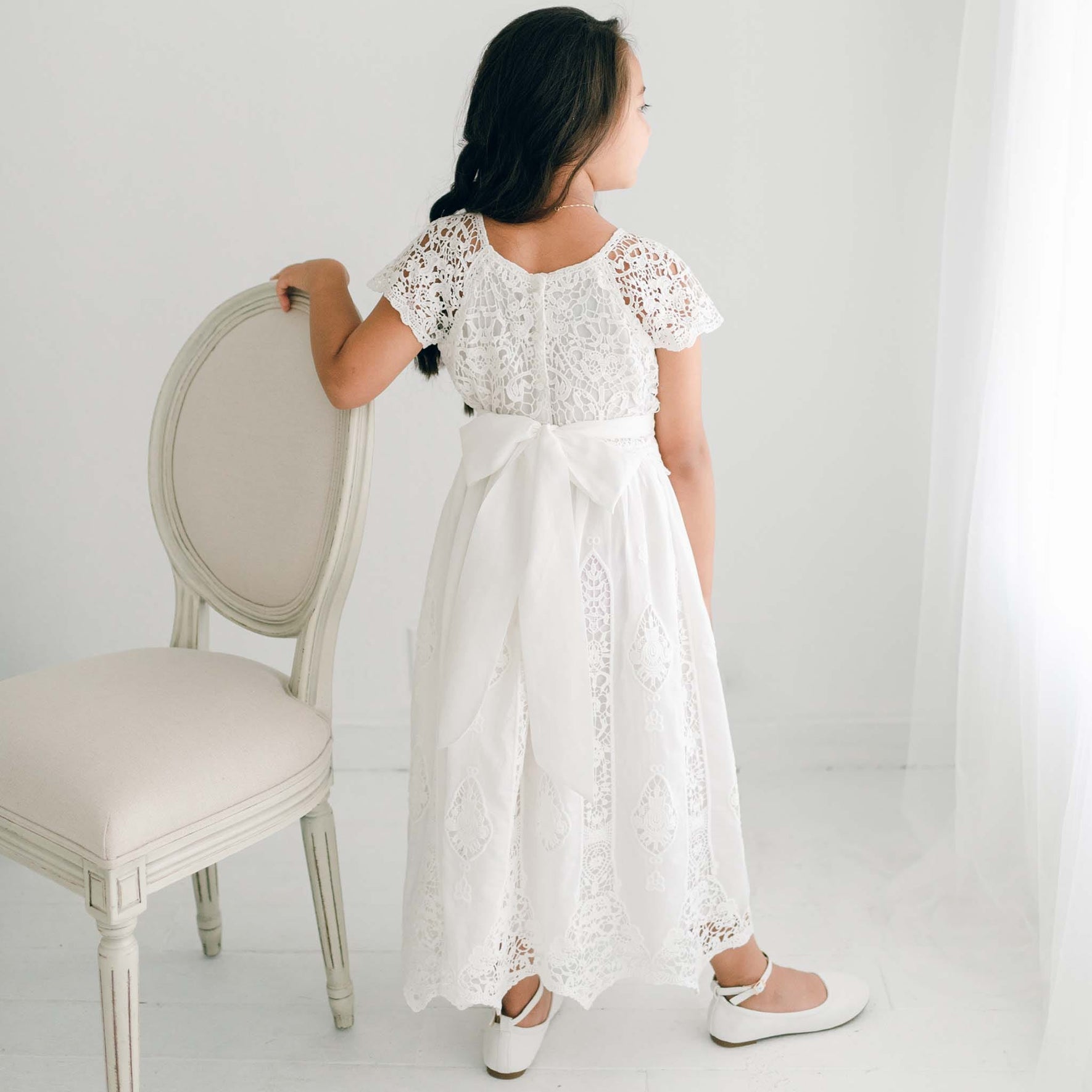 The back view of the Grace Girls Lace Dress, showcasing a wide sash tied into a bow, delicate button closures, and scalloped lace trim.