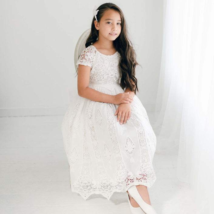 A young girl sits gracefully in the Grace Girls Lace Dress, featuring intricate floral lace patterns and a flowing skirt, as she gazes softly at the camera.