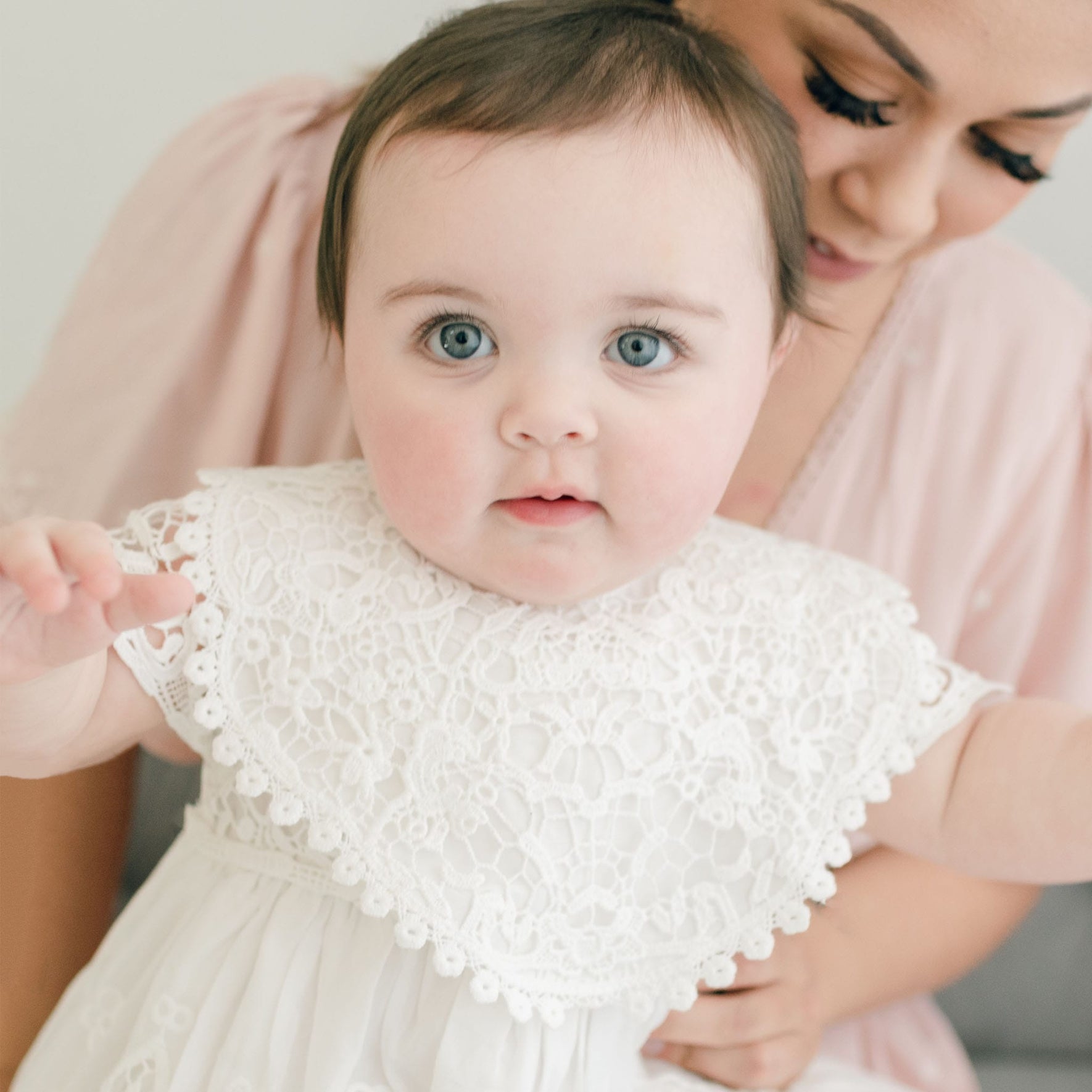A baby with large blue eyes and dark hair, wearing a white christening gown complemented by an intricately designed Grace Lace Christening Bib, is held by an adult in a pink blouse. The adult's face is partially visible, showing a smile and long eyelashes. The baby appears to be reaching forward toward the camera.
