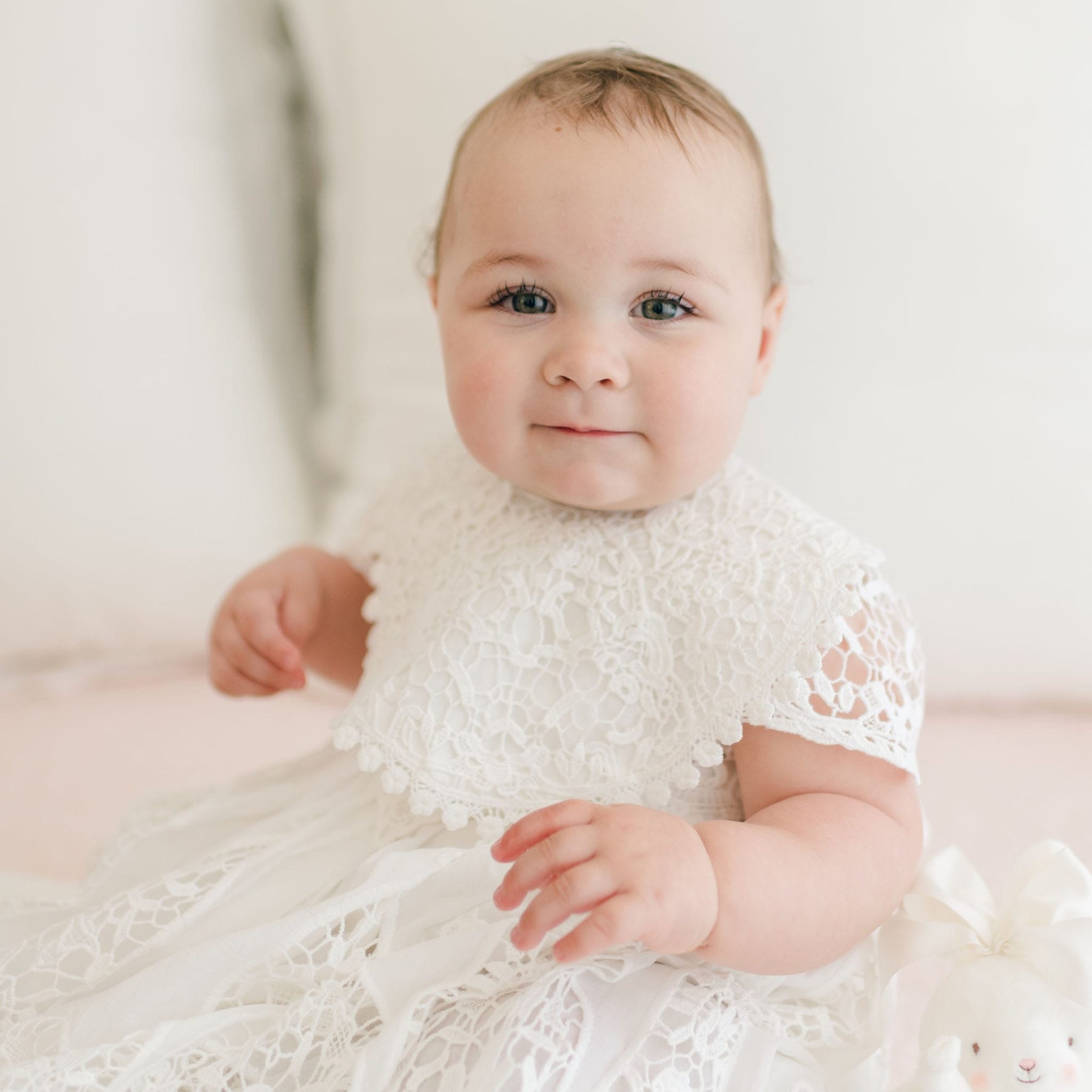 A baby wearing a Grace Lace Christening Bib sits upright, showcasing the intricate lace detailing. The baby has light skin and a slightly smiling expression. In the background, neutral-colored pillows create a soft ambiance, and a small white plush toy is partially visible at the baby's side.